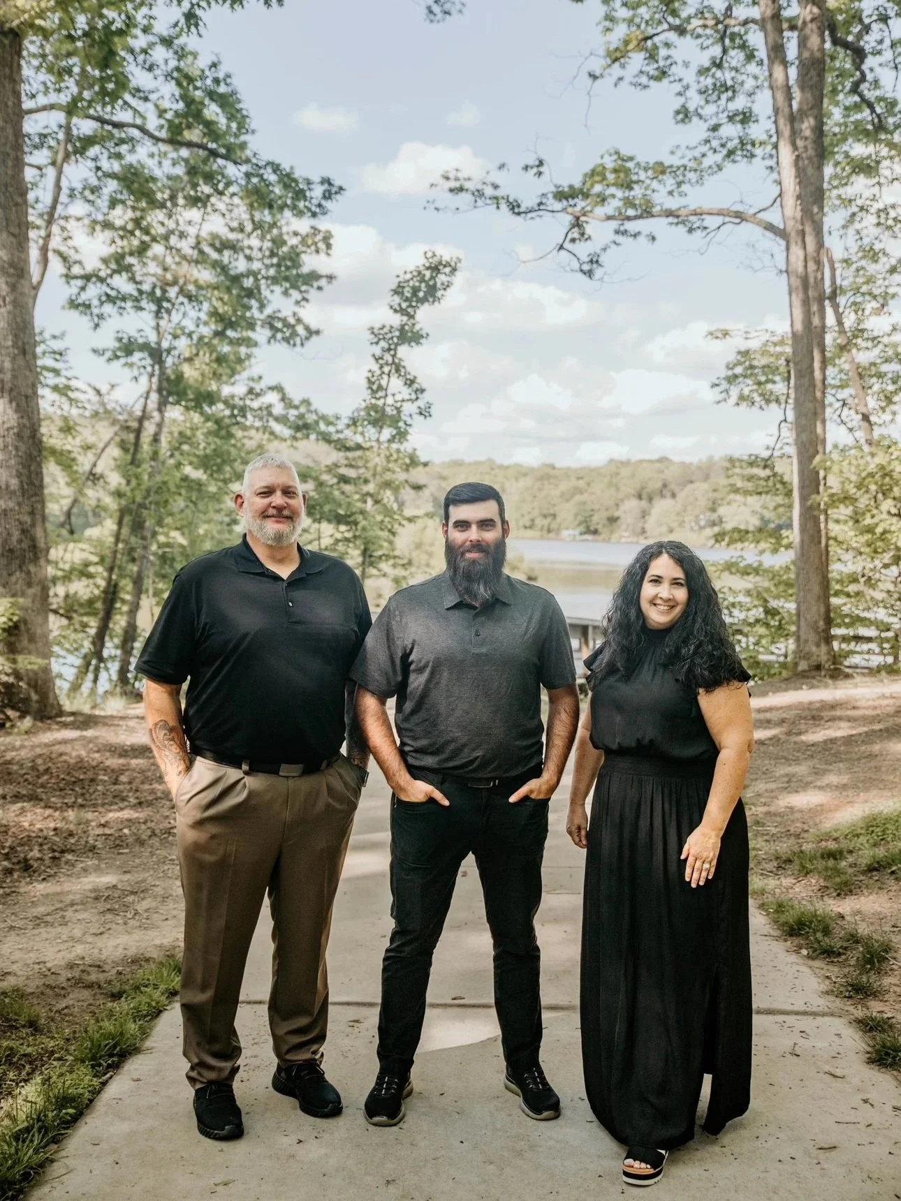 Three people standing outdoors on a sidewalk near trees and water with a partly cloudy sky.