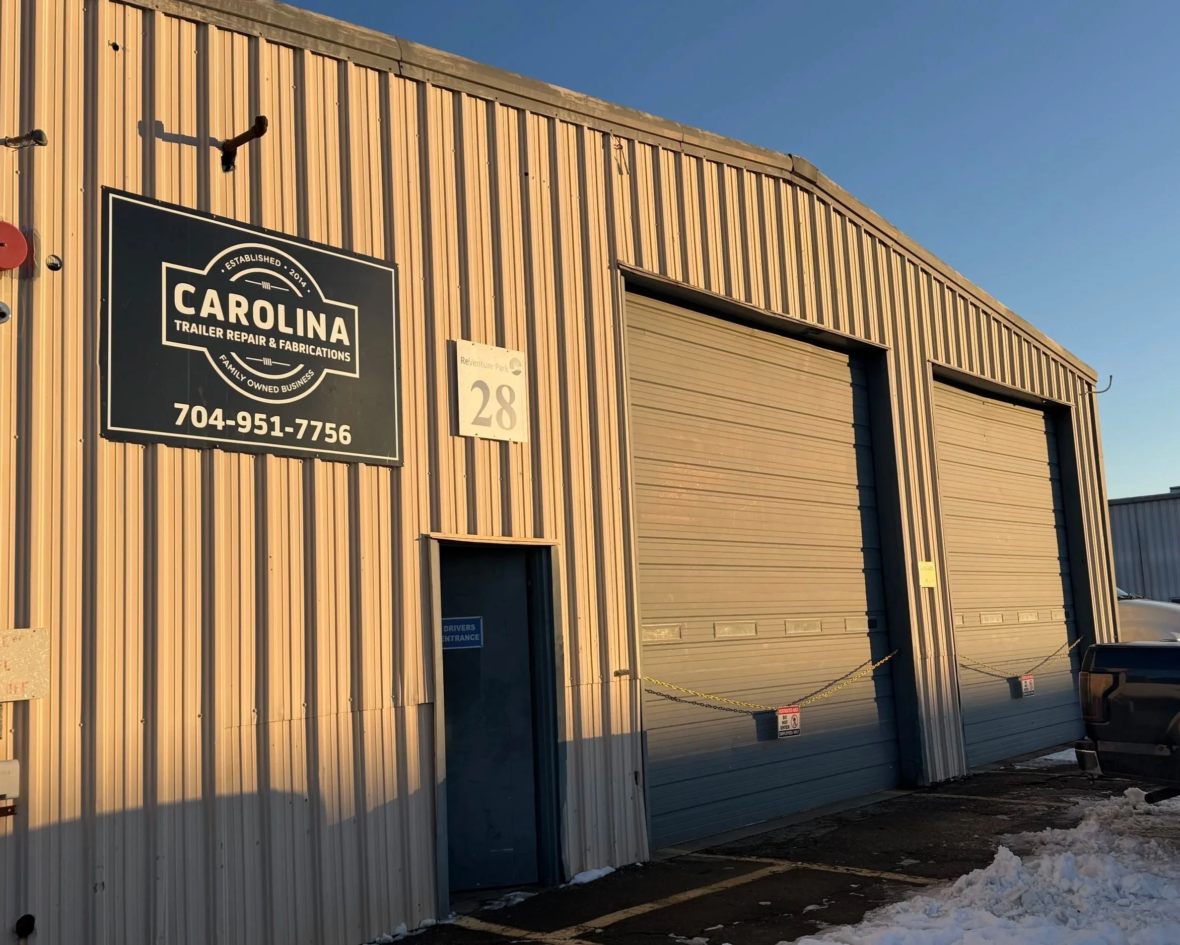 The exterior of a metal building housing Carolina Trailer Repair & Fabrications, a family-owned business established in 2014. The sign displays their phone number 704-951-7756. The building has a large roll-up door, a side door, and a parking area with some snow on the ground.