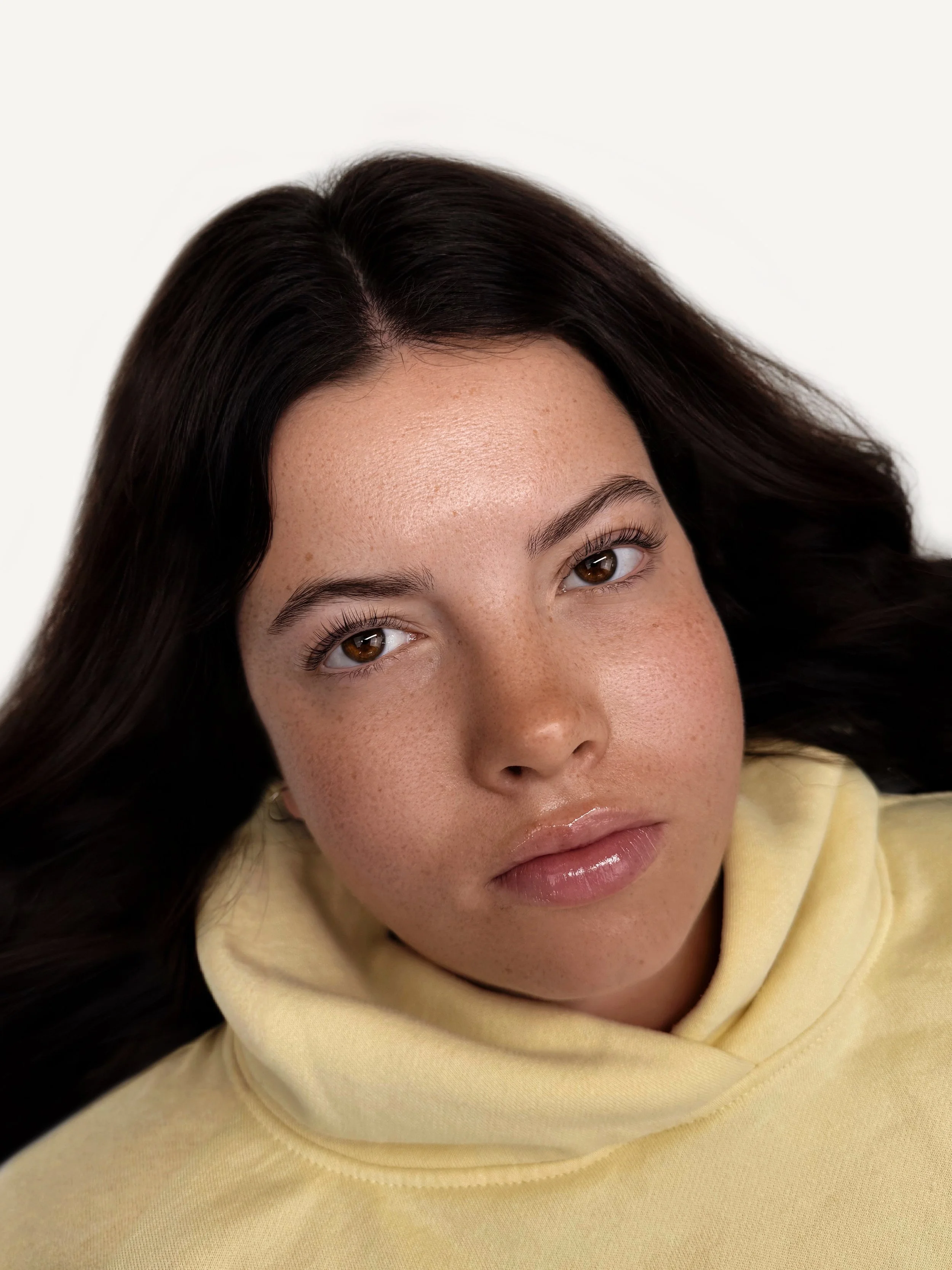 Close-up portrait of a woman with dark hair, light skin with freckles, wearing a yellow hoodie, looking directly at the camera.