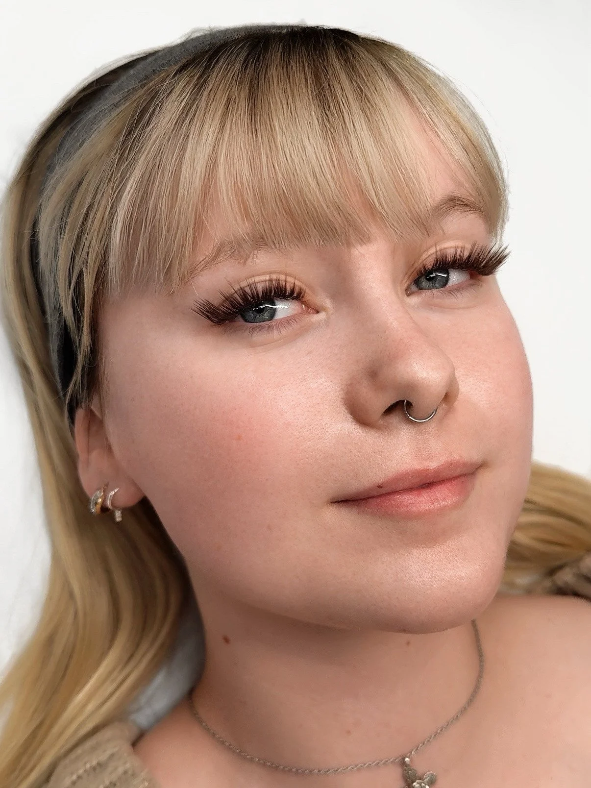 Close-up portrait of a young woman with blonde hair, blue eyes, wearing makeup, a septum piercing, and multiple earrings, with a neutral background.