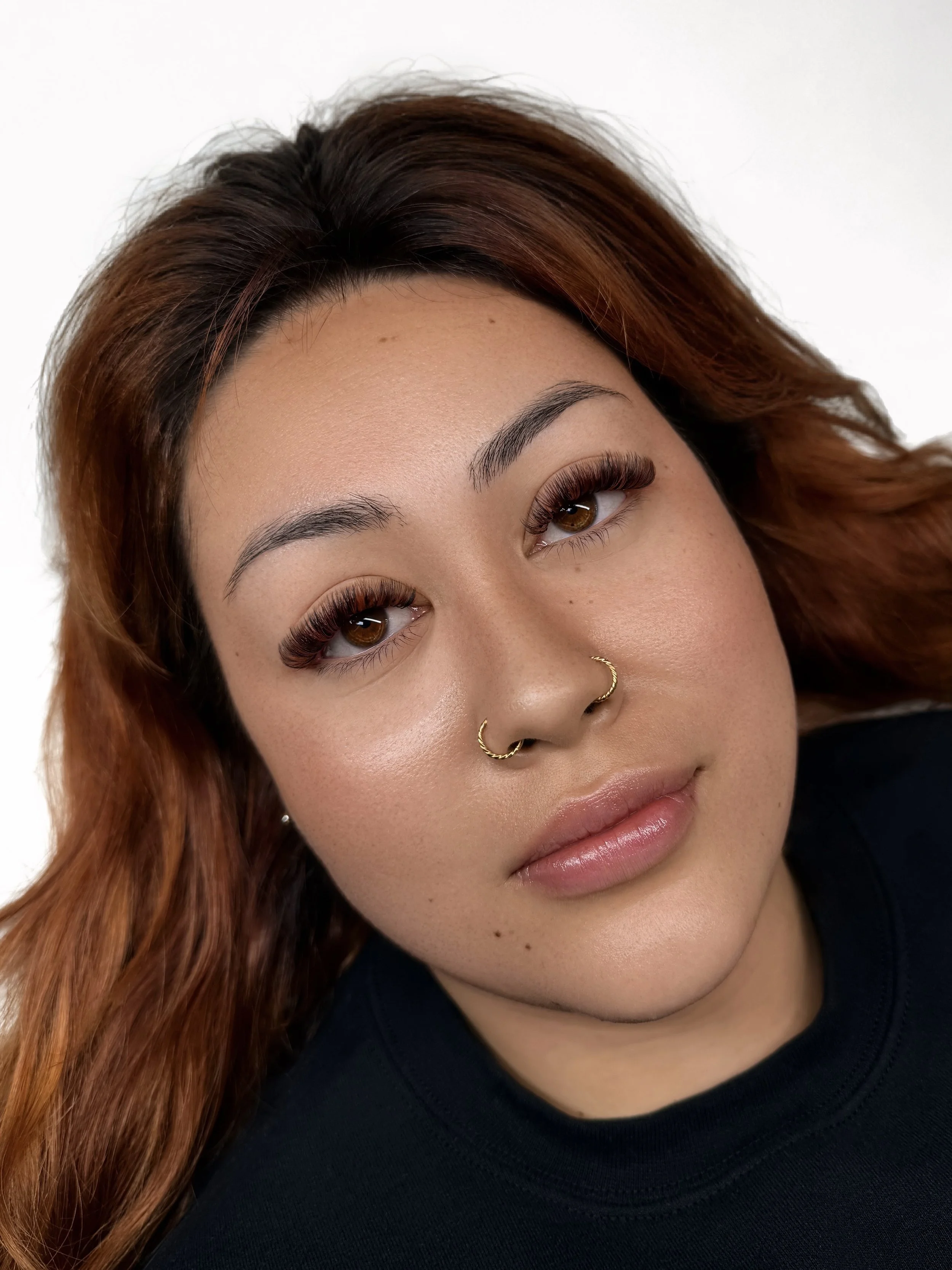 Close-up of a young woman with long brown hair, brown eyes, and makeup, wearing a black top, against a plain white background.