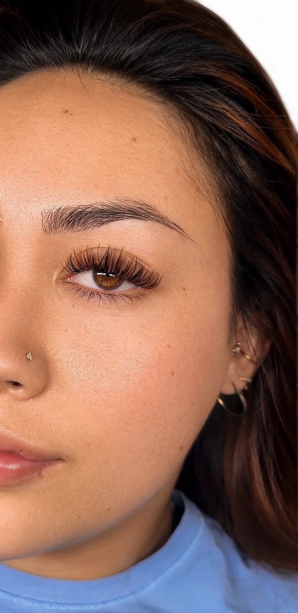 Close-up of a woman's face focusing on the right eye, which has brown irises and long eyelashes. She has a nose piercing on the left nostril, multiple earrings on her right ear, and a natural makeup look with clear skin and lightly filled eyebrows.