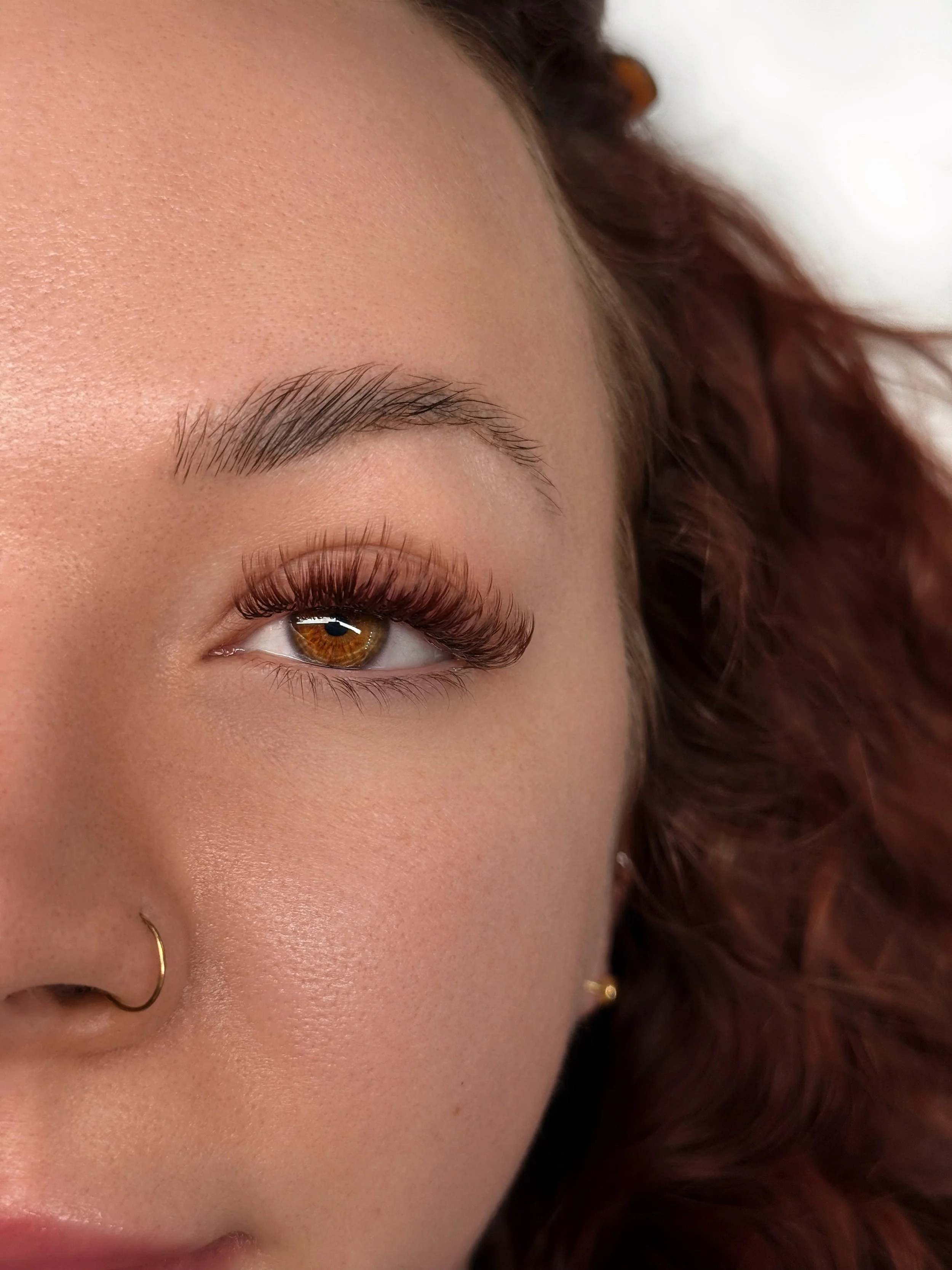 Close-up of a woman's face, focusing on her eye with long eyelash extensions, her eyebrow, and part of her nose with a gold nose ring. She has fair skin and wavy auburn hair.