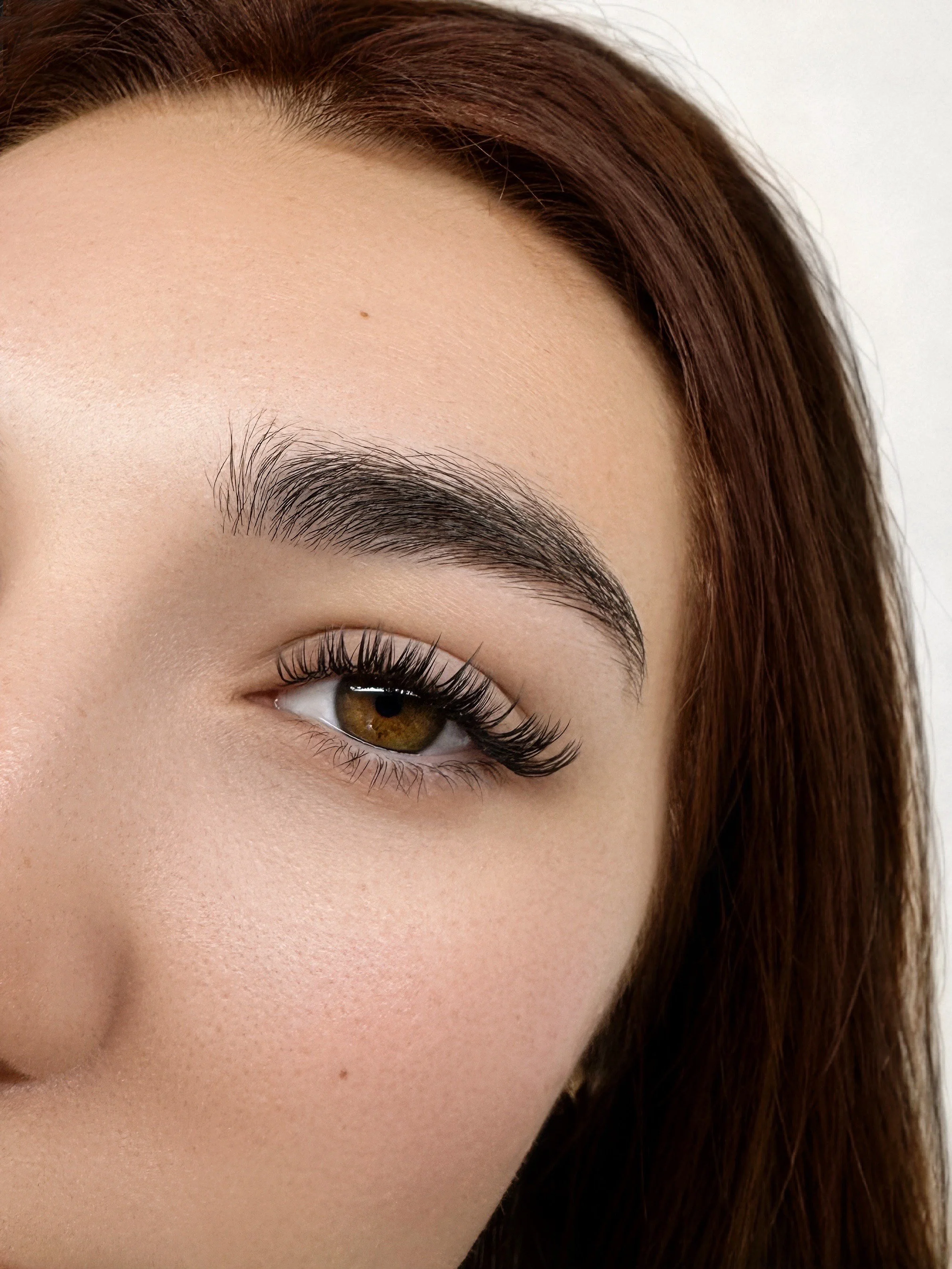 Close-up of a woman's eye with long eyelashes, well-defined eyebrow, and smooth skin with a small mole and freckles, with brown hair partially visible.