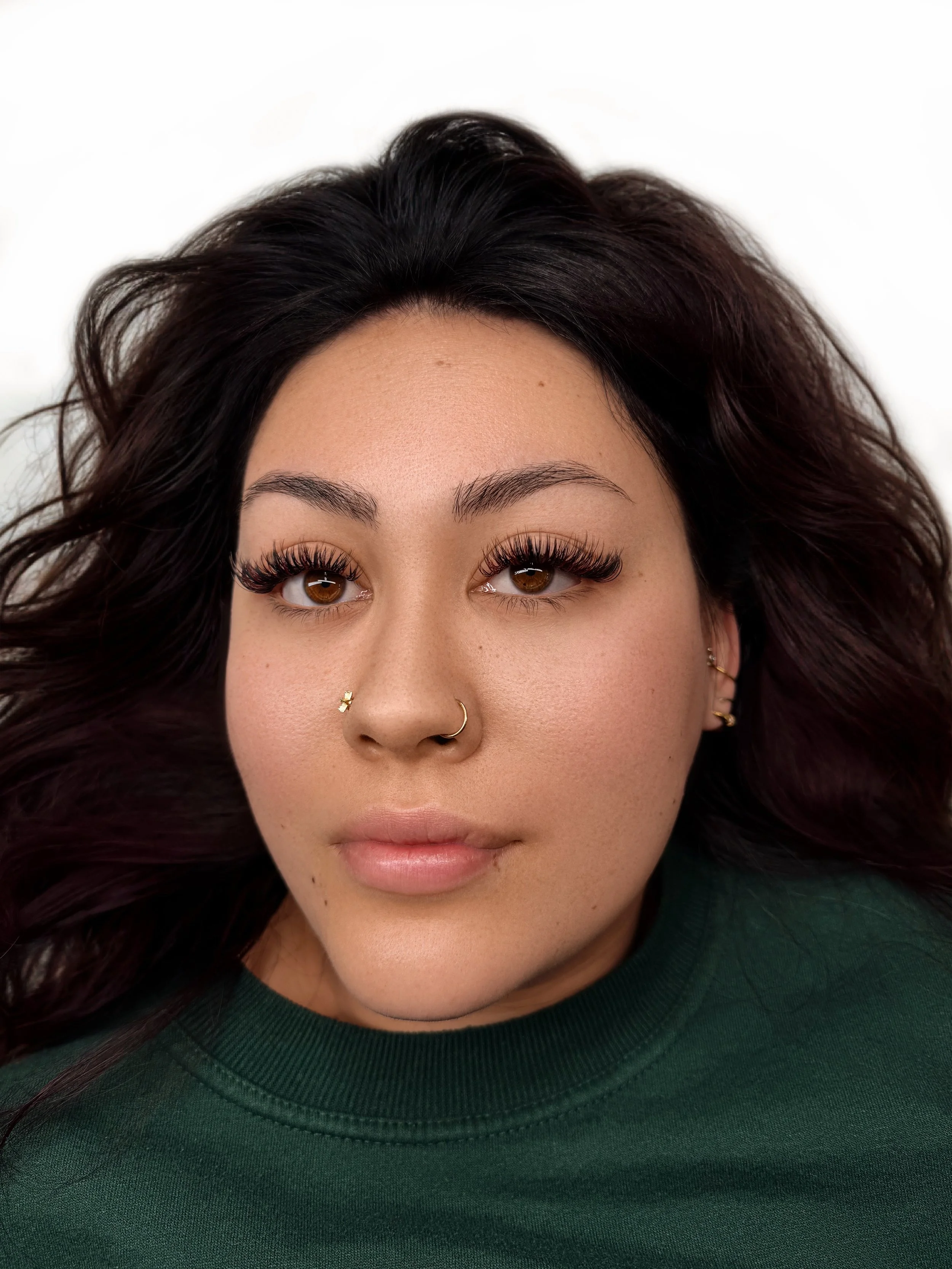 Close-up of a woman with long dark wavy hair, brown eyes, wearing makeup, a nose ring, and multiple earrings, wearing a green shirt, against a white background.