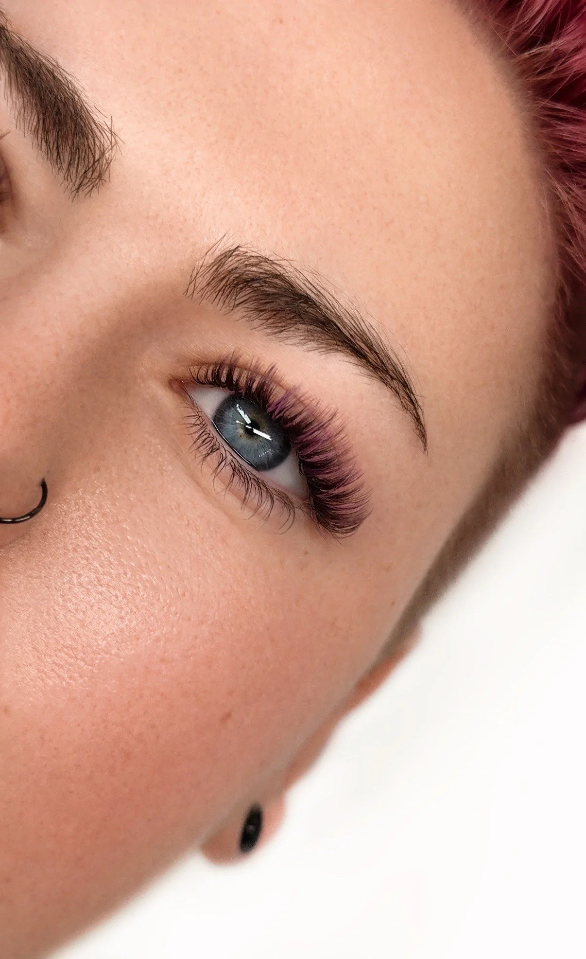 Close-up of a person's face showing their eye, eyebrow, and part of their nose with a septum piercing. The eye has makeup with long lashes and a hint of purple eyeshadow, and the person has pink hair.