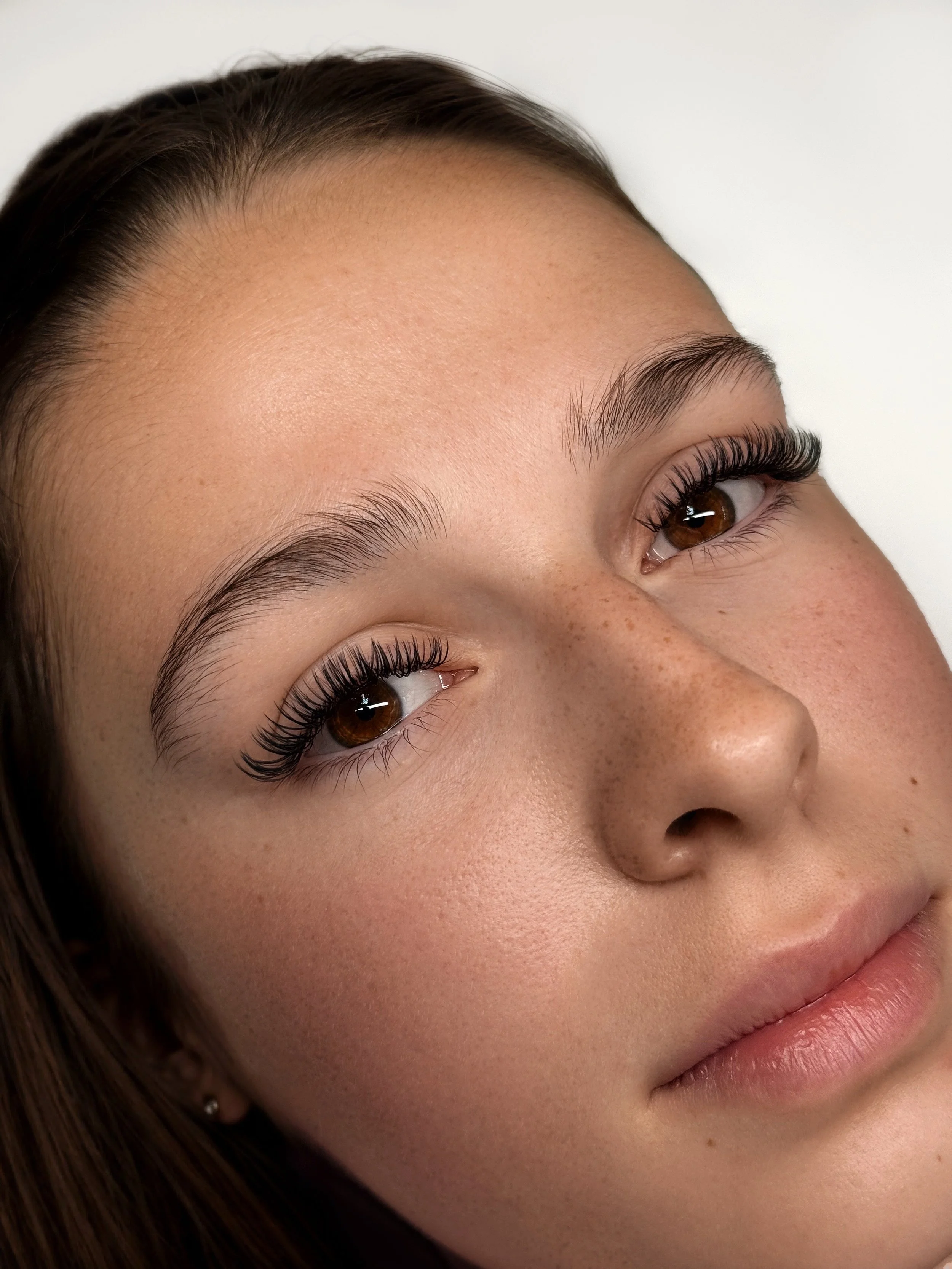 Close-up of a young woman's face with brown eyes, natural eyebrows, and long eyelashes, against a plain white background.