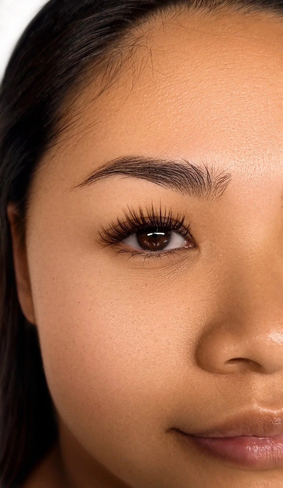 Close-up of a woman's face showing her eye, eyebrow, part of her nose, and lips with makeup and clear skin.
