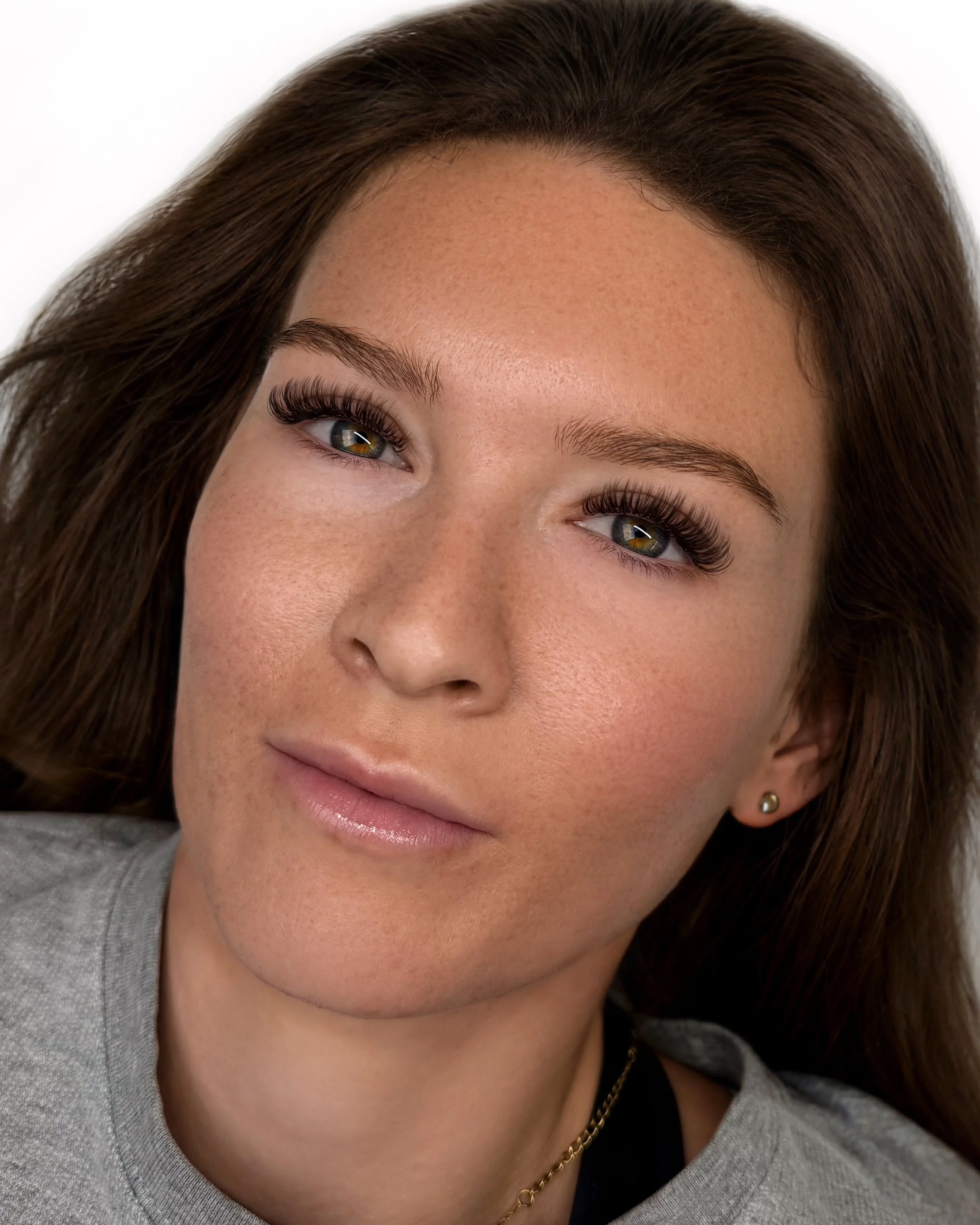 Close-up portrait of a woman with brown hair, green eyes, natural makeup, and a light gray top, looking directly at the camera.