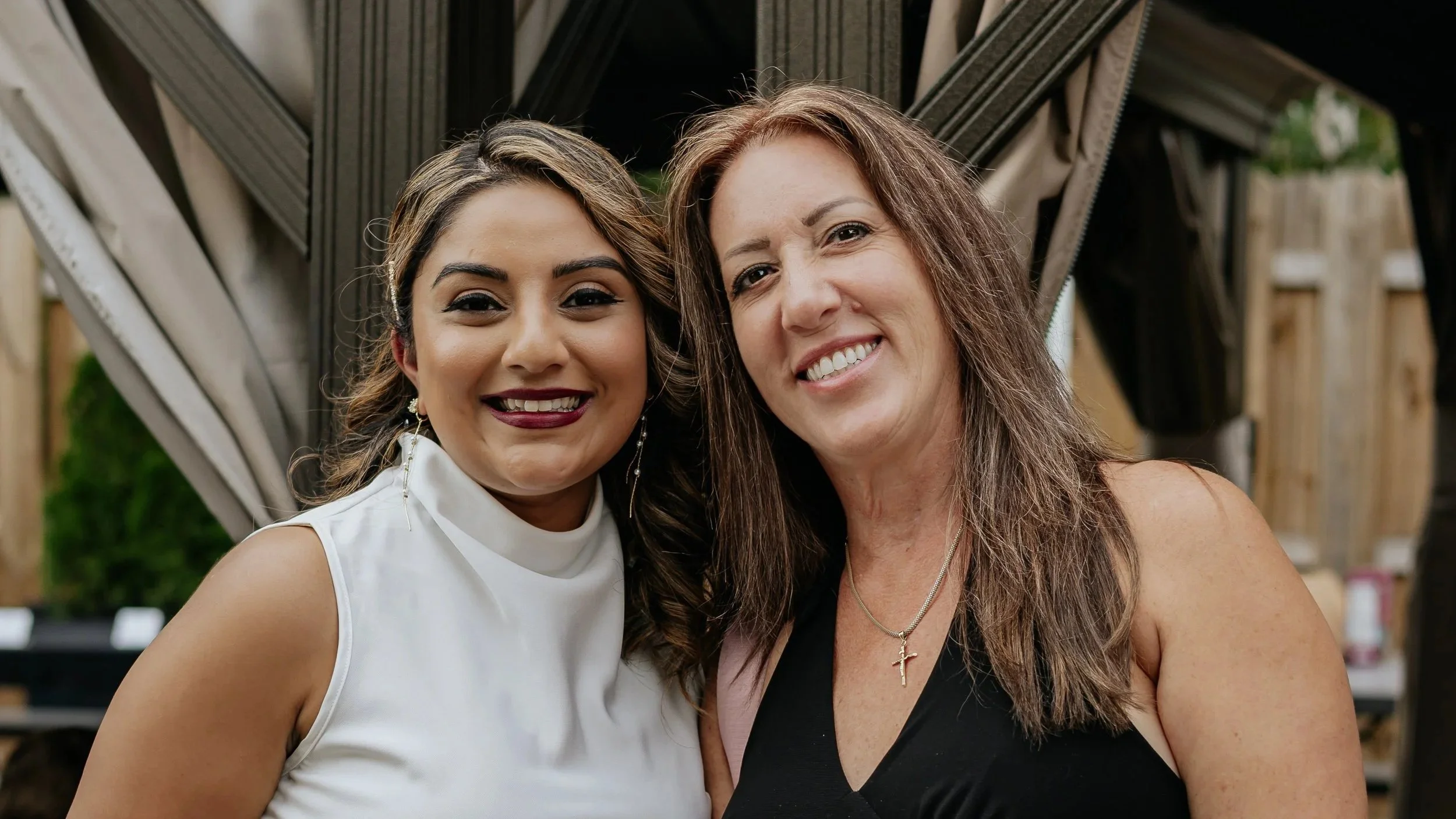 Two women smiling closely together outdoors, with a wooden fence and garden in the background.