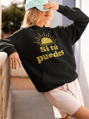 Person wearing a black sweatshirt with yellow text that says "Sí tú puedes," and a light blue cap, standing outdoors.