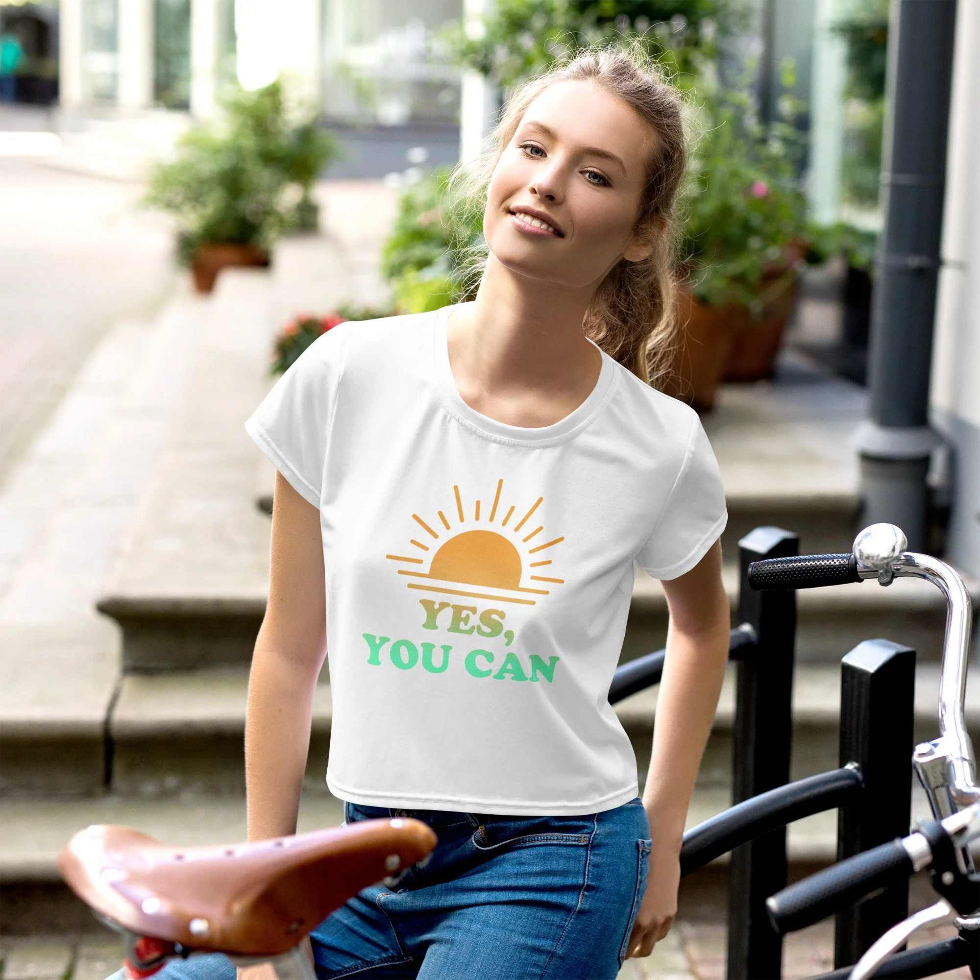 A young woman standing outdoors on a sidewalk, next to a bicycle, wearing a white T-shirt with a sunrise graphic and the words 'Yes, You Can'.