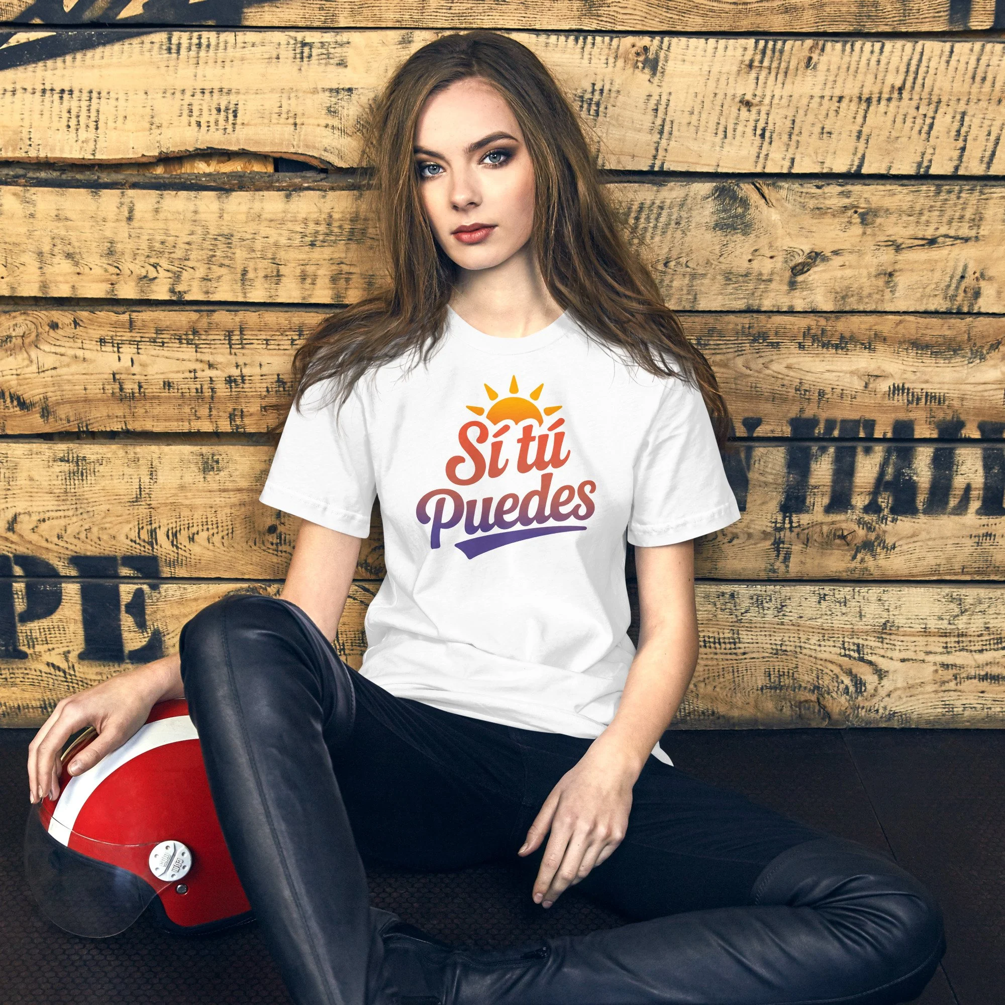 A young woman with long brown hair and blue eyes, sitting against a wooden wall, wearing a white t-shirt with the phrase 'Si tú Puedes' and a sun graphic, black leather pants, and holding a red motorcycle helmet.