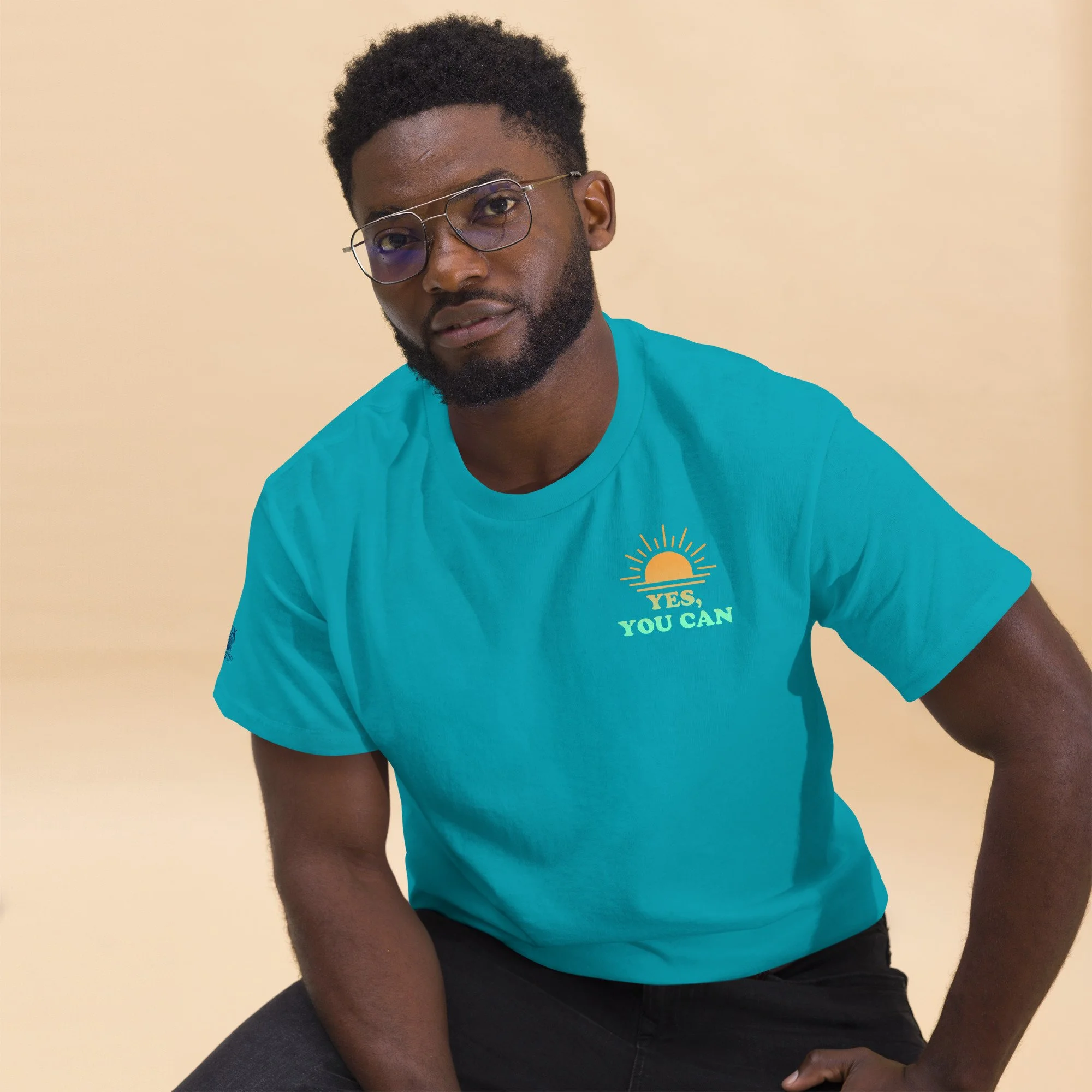 A young man wearing glasses and a turquoise t-shirt with a sun graphic and the text 'YES, YOU CAN'. he has a beard and is sitting against a beige background.