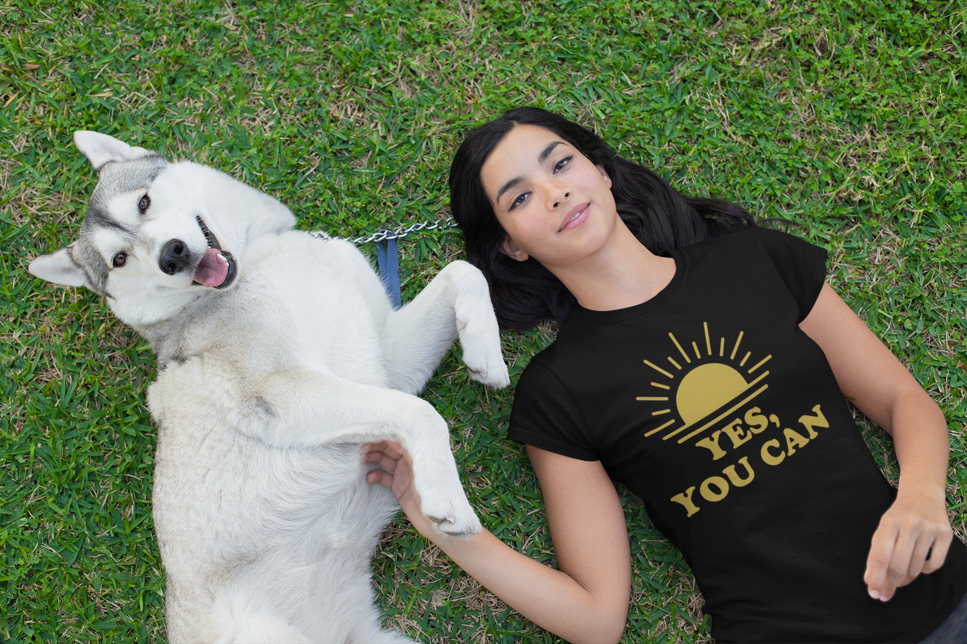 Woman lying on grass in black ‘Yes, You Can’ T-shirt with sun graphic, playing with Siberian Husky — joyful lifestyle fashion from E.N.K. Feel Good Apparel