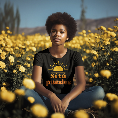 A young woman with curly hair sitting cross-legged in a field of yellow flowers, wearing a black T-shirt with an orange sun and the words 'Sí tú puedes'.