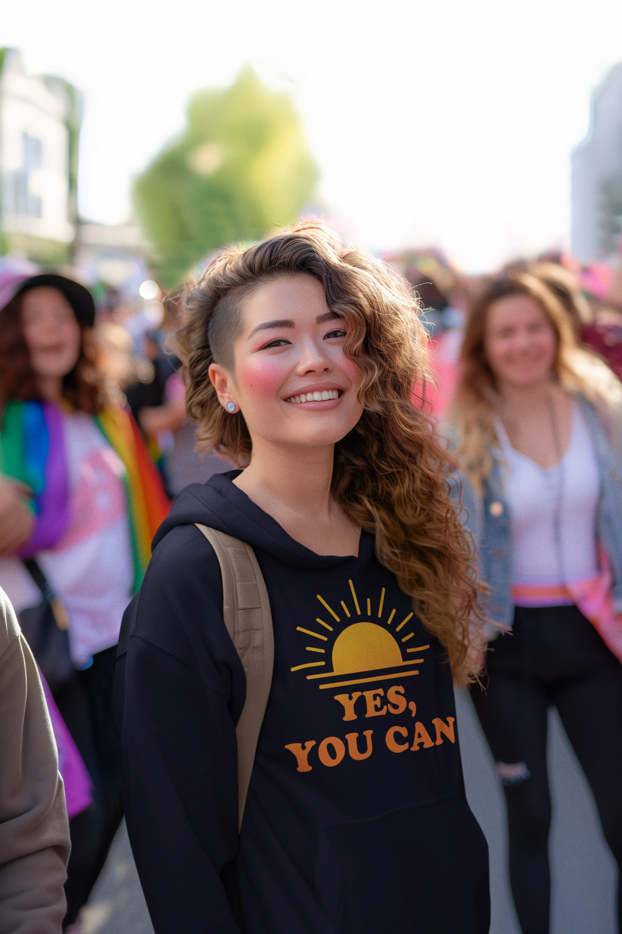 Young woman with wavy hair smiling wearing a black hoodie that says "Yes, you can" with a sunset graphic, standing outdoors with a group of people in the background at a public event.