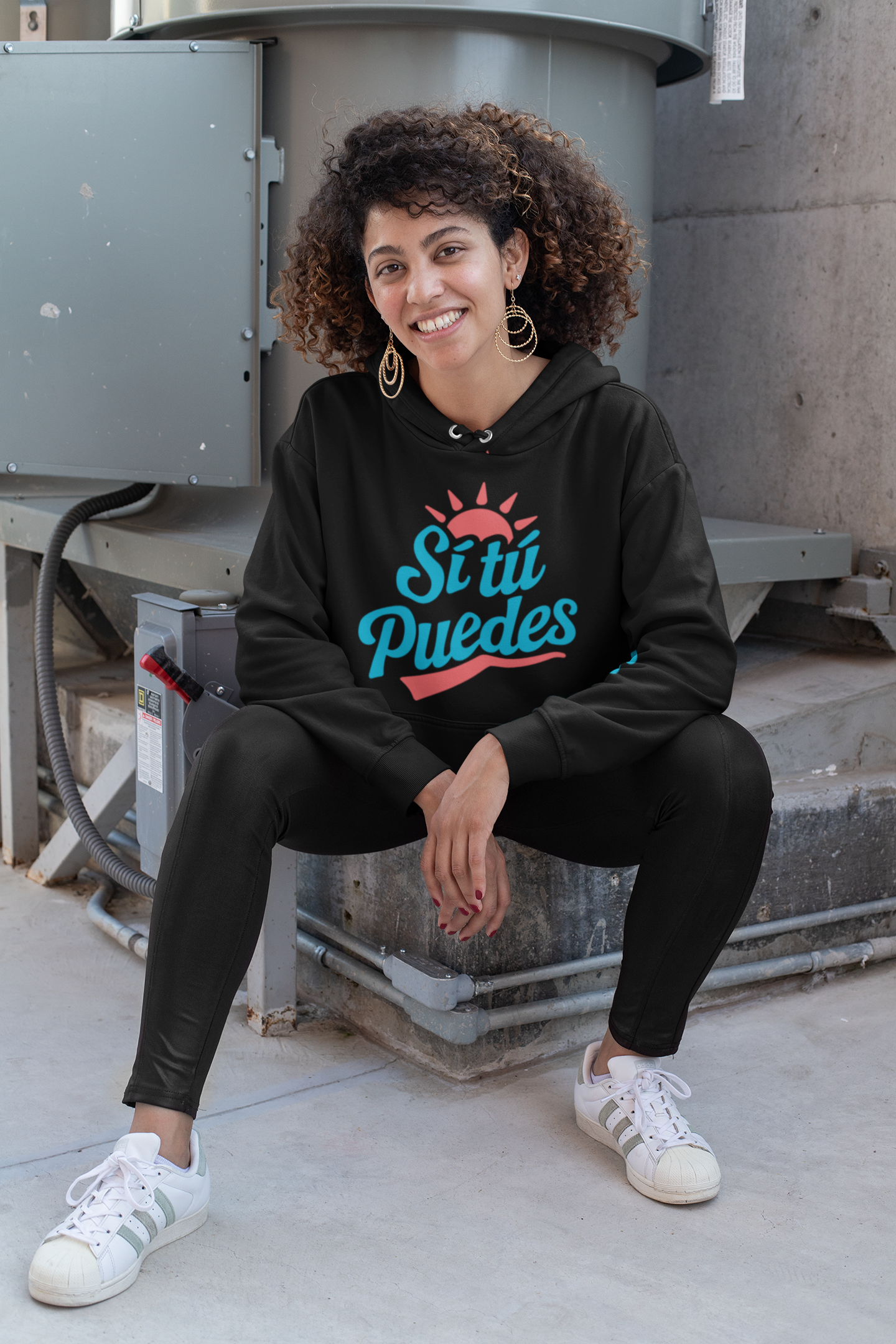 Curly-haired woman wearing black ‘Sí Tú Puedes’ hoodie with sun graphic, relaxed outdoor street style — E.N.K. Feel Good Apparel