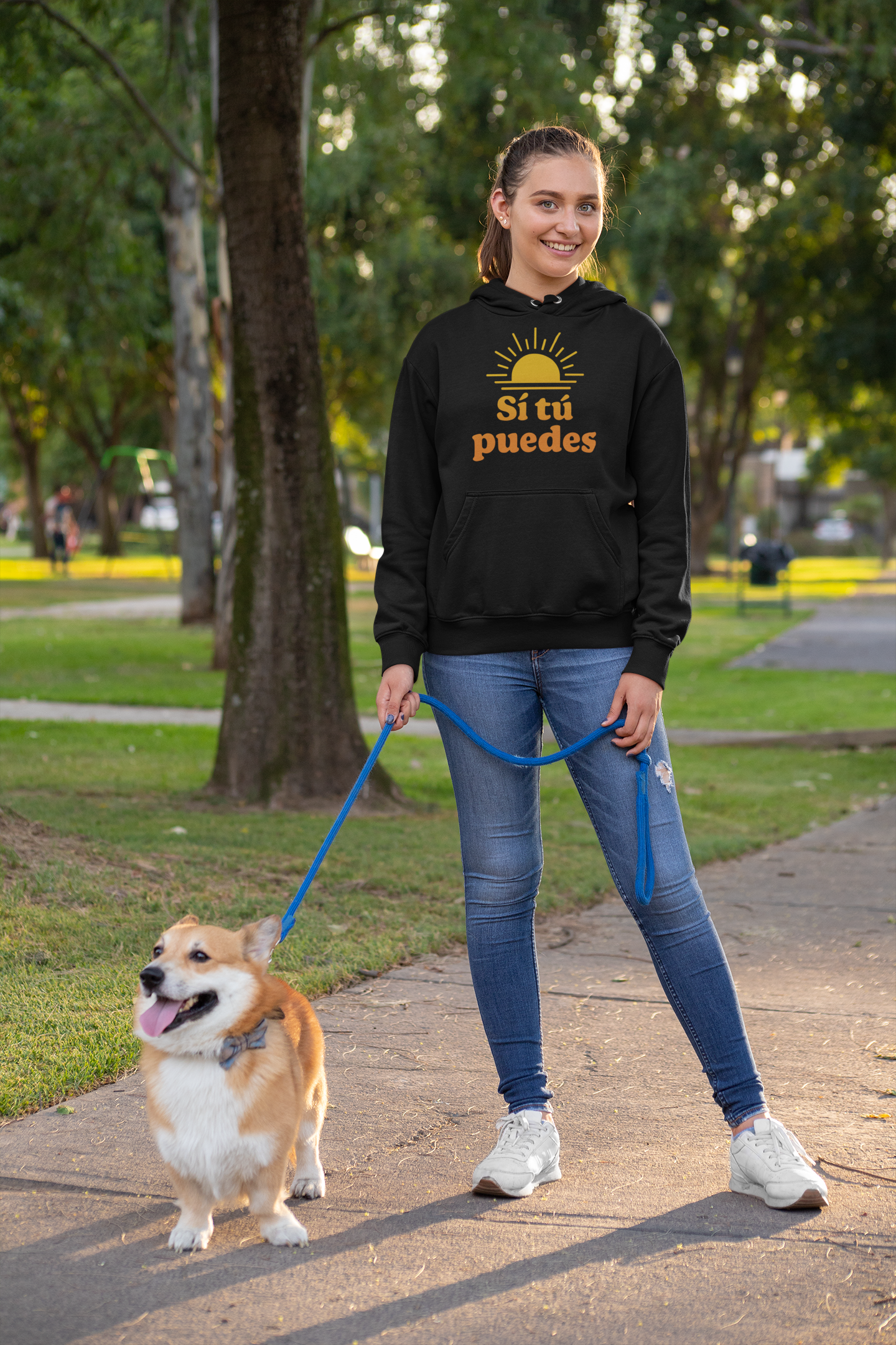 A young woman in a black hoodie with the words 'Sí tú puedes' and a sun graphic, smiling and holding a blue leash attached to a small, fluffy Corgi dog walking on a paved path in a park with green trees.
