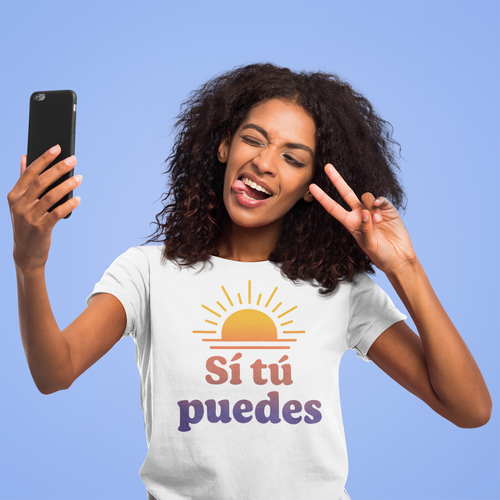 A young woman with curly hair taking a selfie, making a peace sign with her fingers, wearing a white T-shirt with a sun graphic and the Spanish phrase 'Sí tú puedes' against a blue background.
