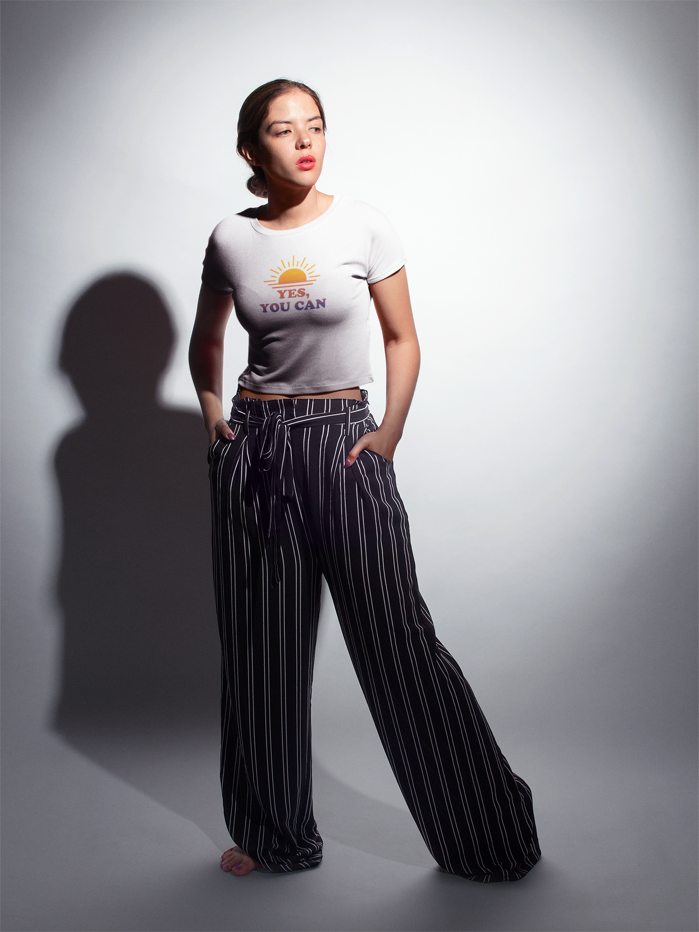 A young woman with brown hair tied back, wearing a white crop top with a sunset graphic and the words 'YES, YOU CAN' printed on it, black and white striped wide-leg pants, standing barefoot against a plain white background with a shadow cast to her l
