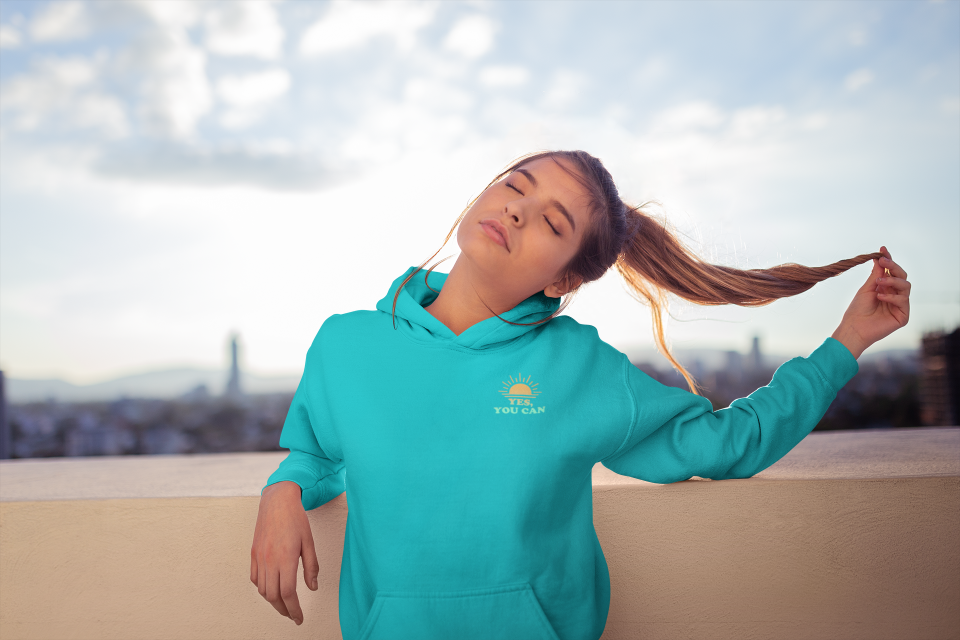 Smiling woman in turquoise ‘Sí Tú Puedes’ hoodie with sun design, standing on rooftop in sunlight — uplifting urban style by E.N.K. Feel Good Apparel
