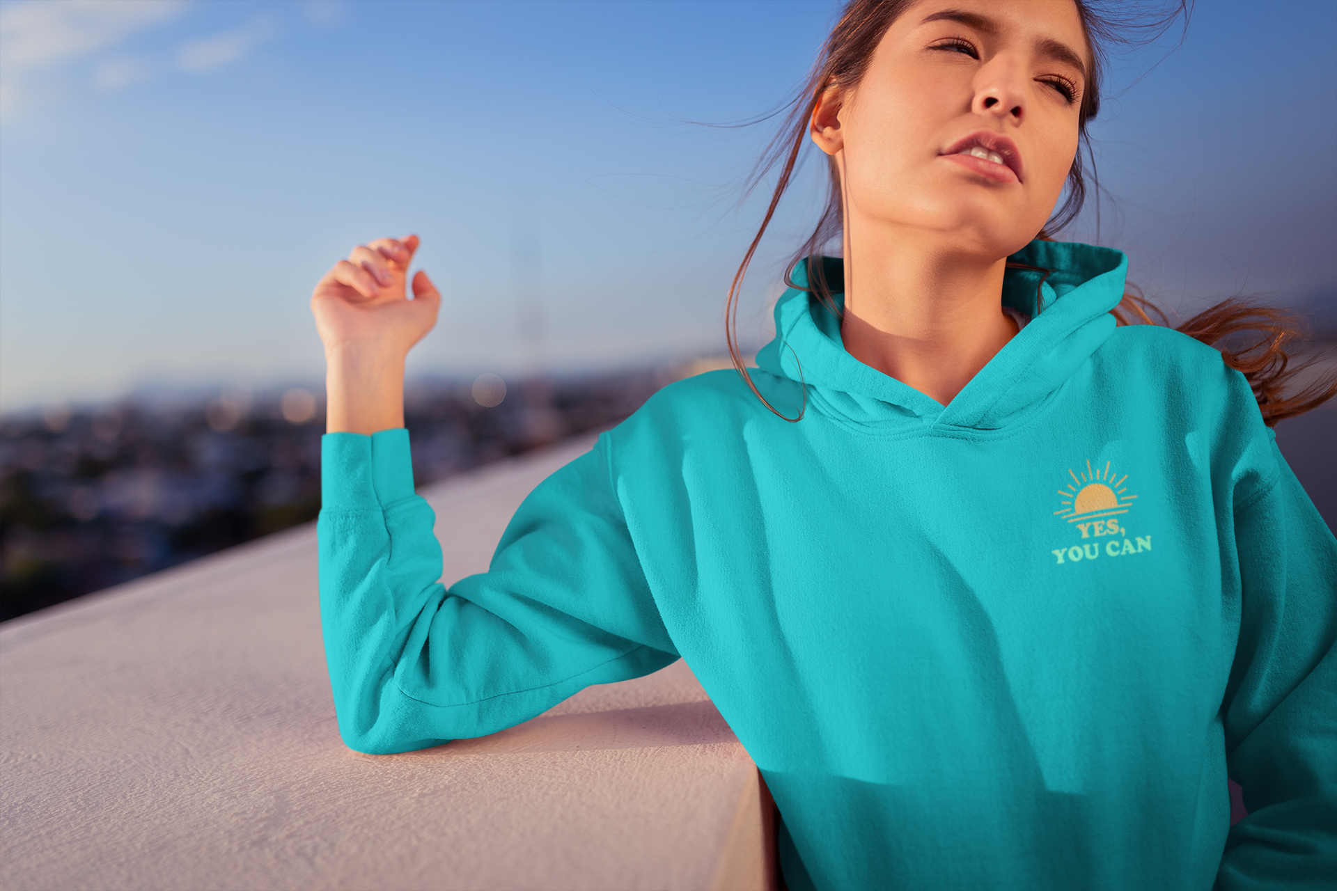 Young woman with brown hair wearing a teal hoodie with a sunrise and the text 'YES, YOU CAN' on an outdoor rooftop or balcony during sunset.