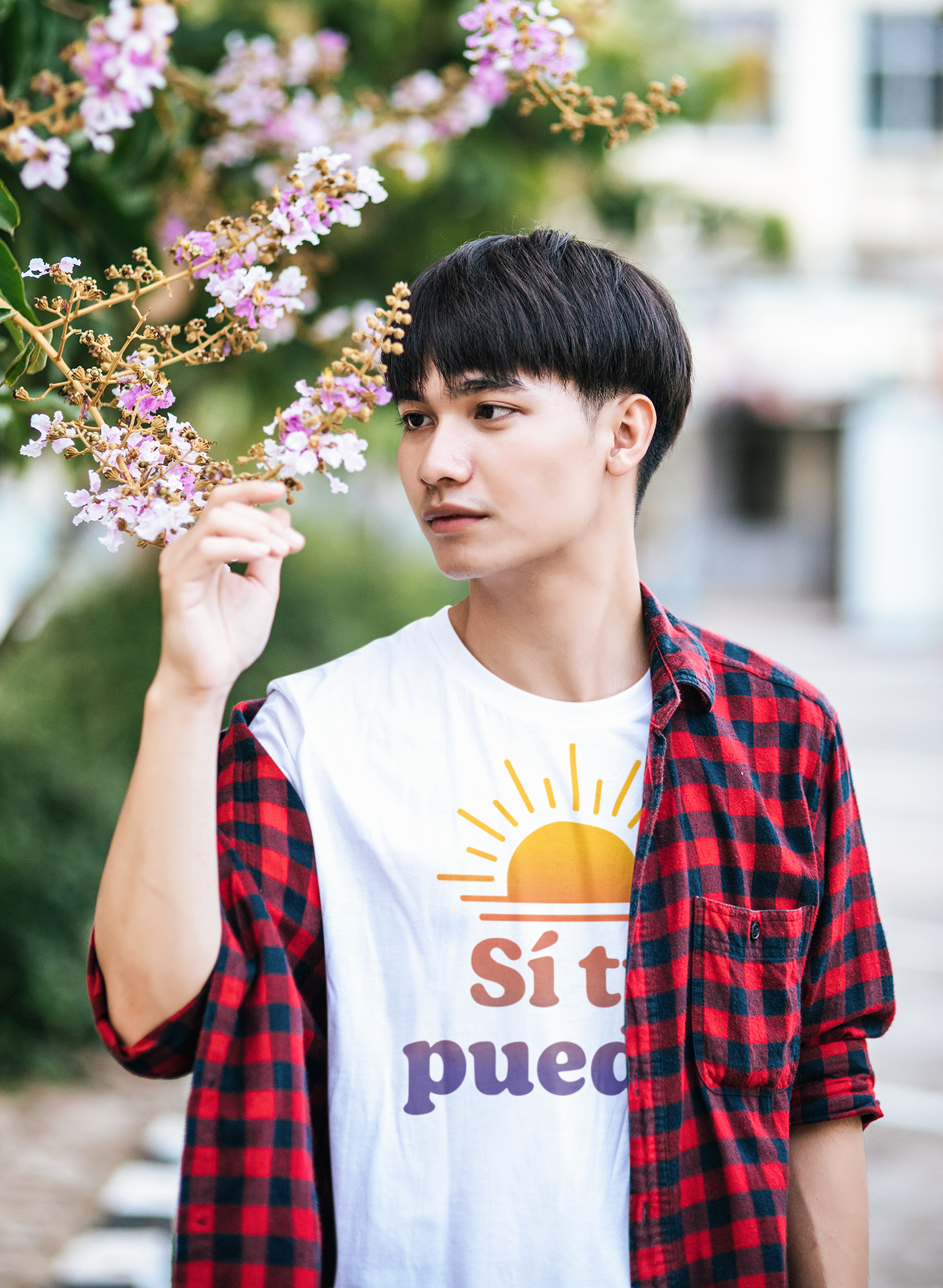 Young man wearing white ‘Sí Tú Puedes’ T-shirt with sunset graphic, layered with plaid shirt, outdoors near flowers — casual motivational style from E.N.K. Feel Good Apparel.