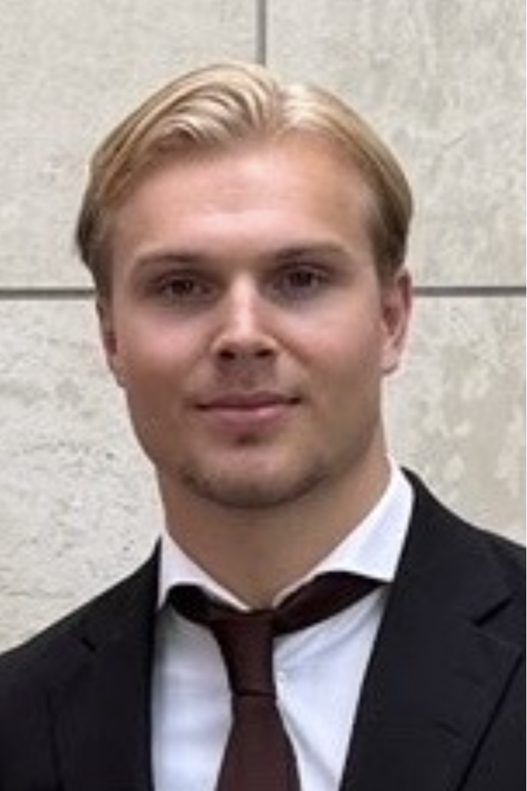 En ung man i kostym och slips står framför en ljus vägg.