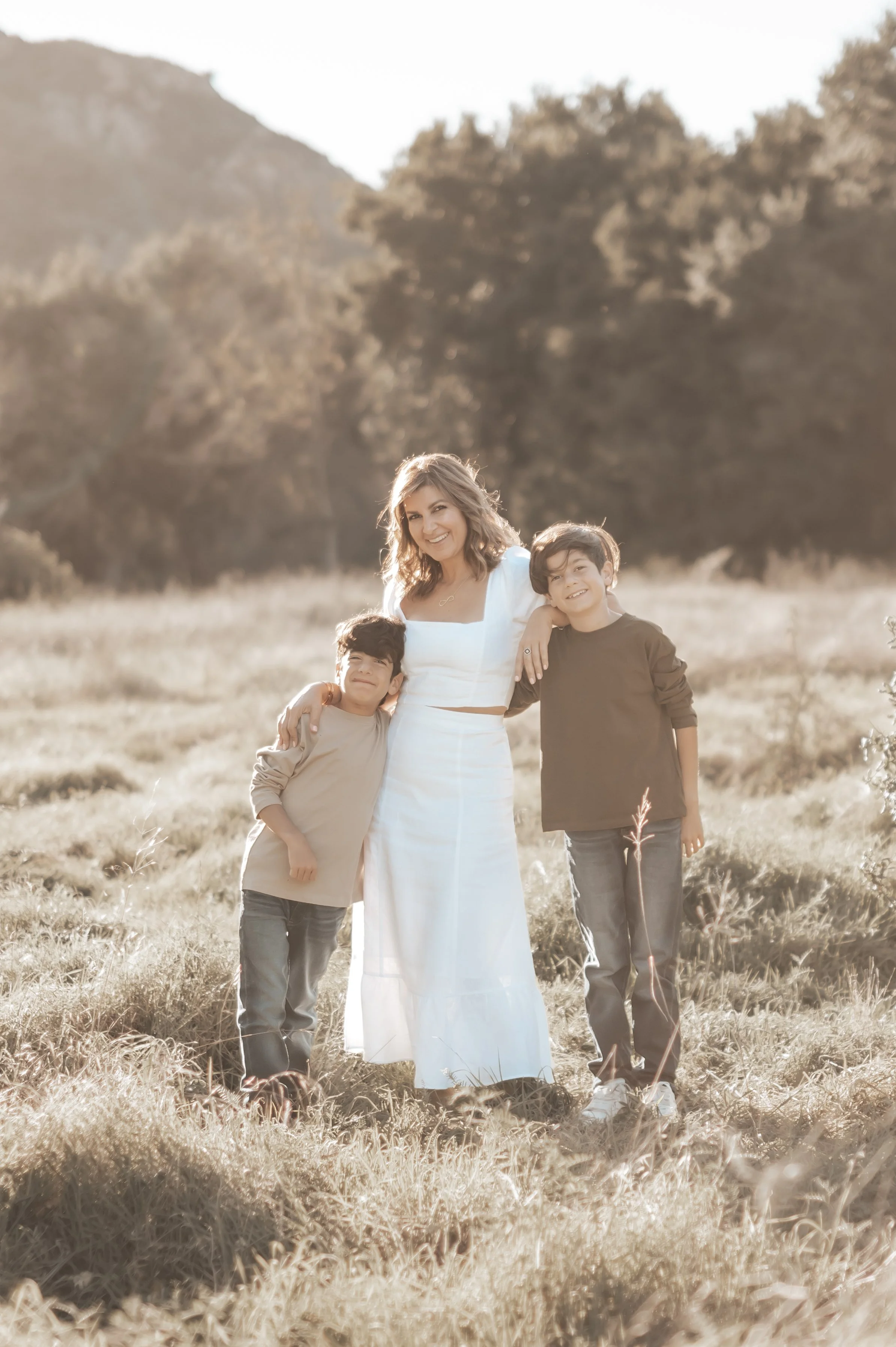 A woman and two boys standing outdoors in a grassy field with mountains in the background, smiling