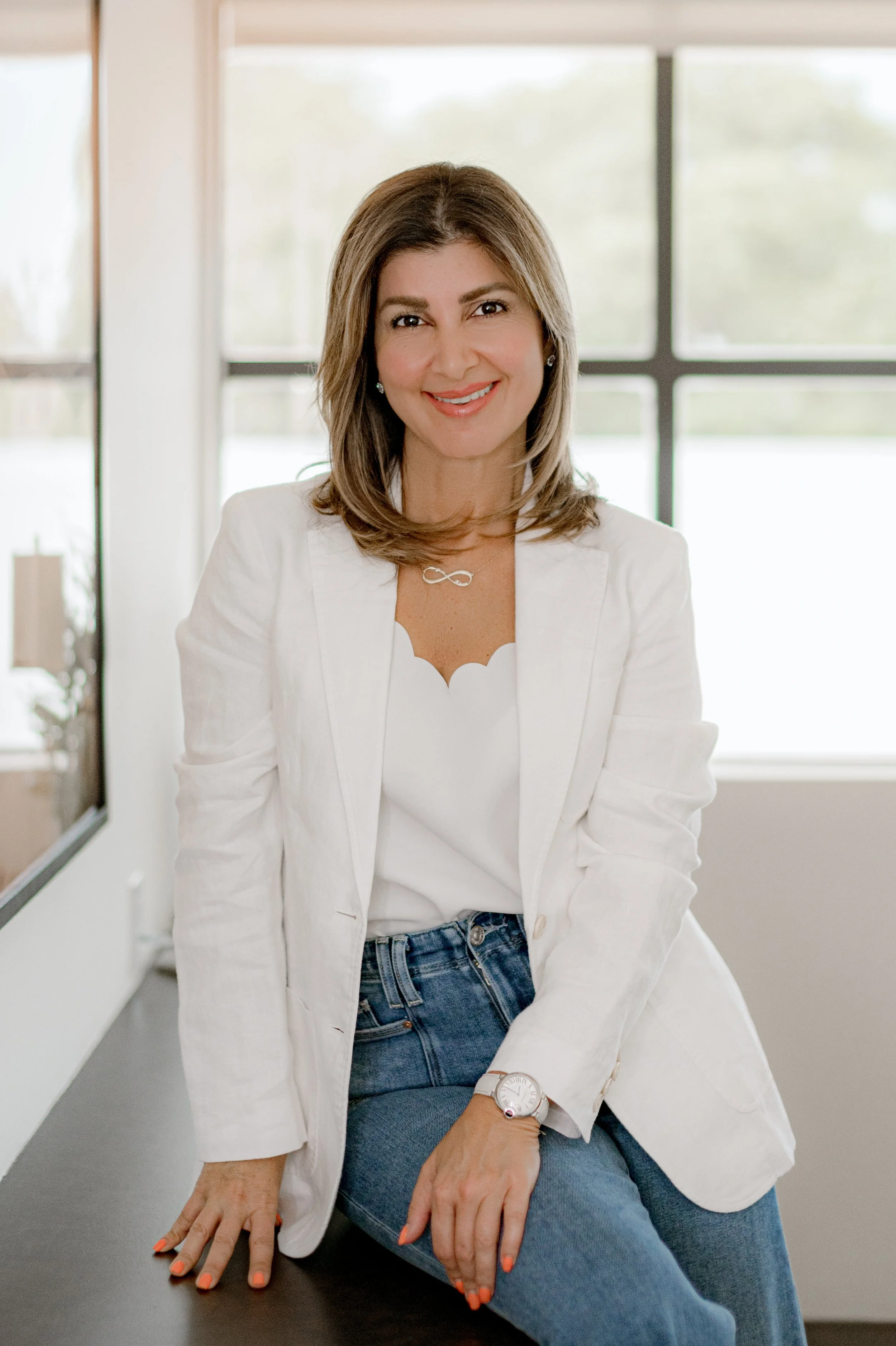 A woman with shoulder-length brown hair, wearing a white blazer and jeans, sitting on a black counter in a bright room with large windows and smiling.