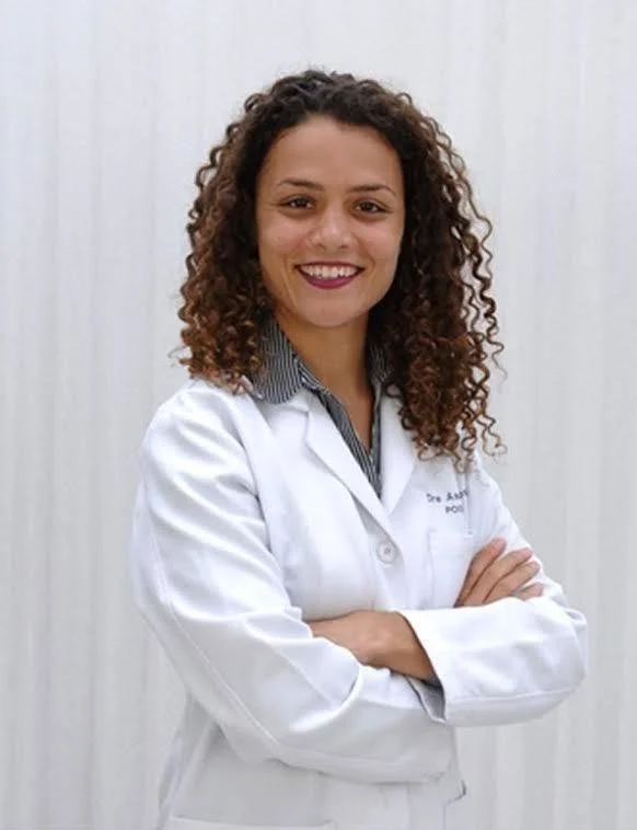 A podiatrist with curly brown hair wearing a white lab coat and standing with arms crossed against a plain background.