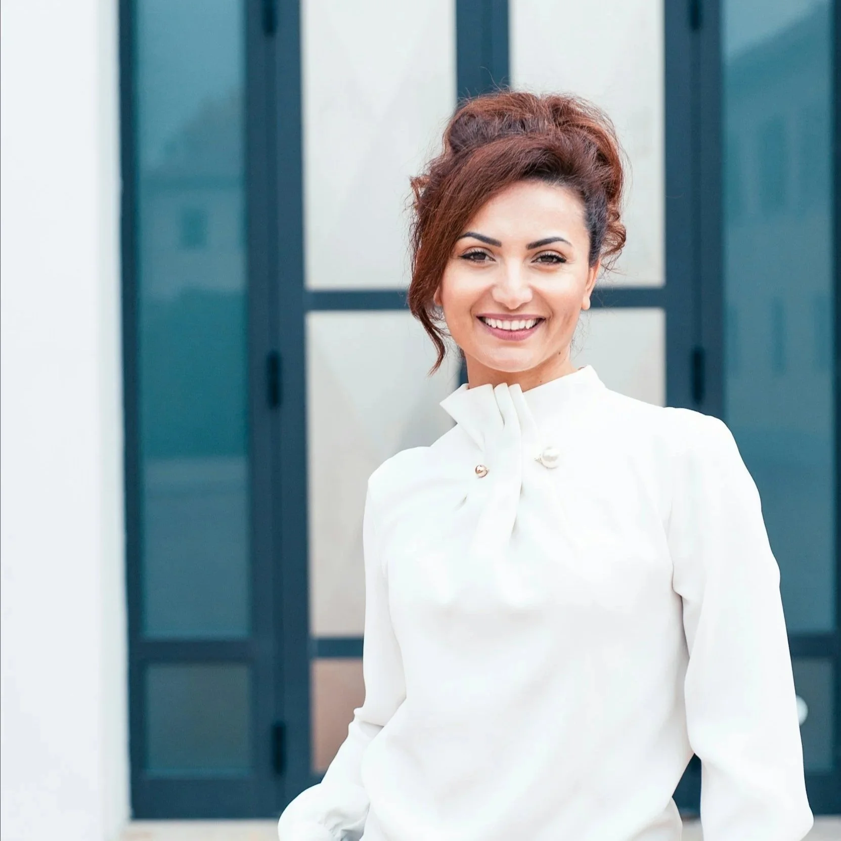 A smiling woman with dark hair styled in an updo, wearing a white blouse with a bow at the collar, standing outdoors in front of a building with glass doors.