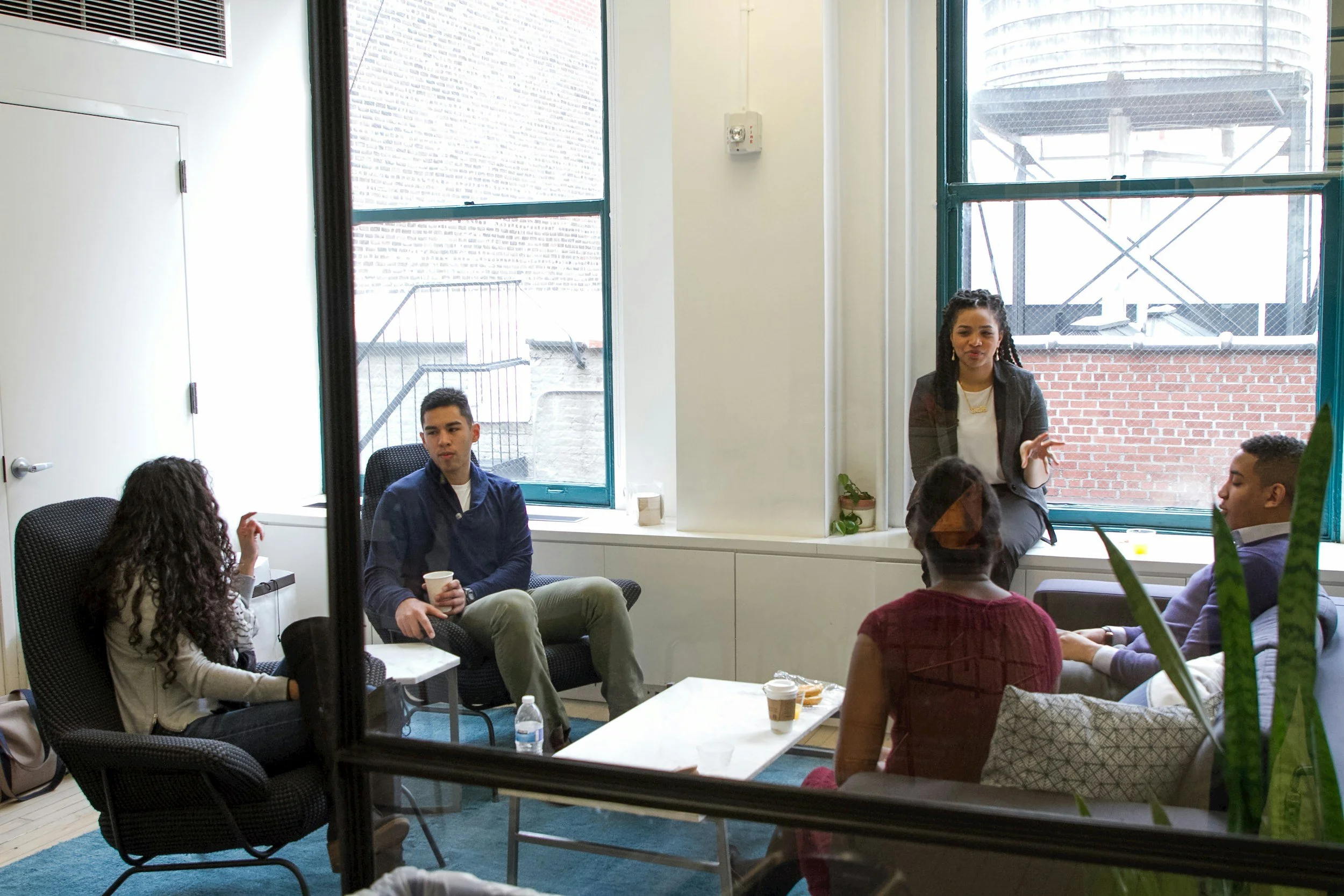 A diverse group of six young adults participating in a discussion in a brightly lit, casual office or meeting room, with large windows showing an urban brick building outside.