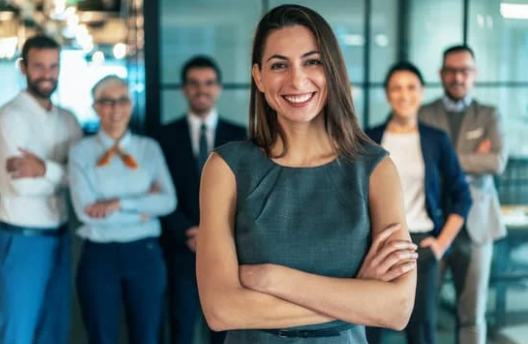 A confident woman smiling with her arms crossed, standing in front of a diverse group of five professionally dressed coworkers in a modern office.