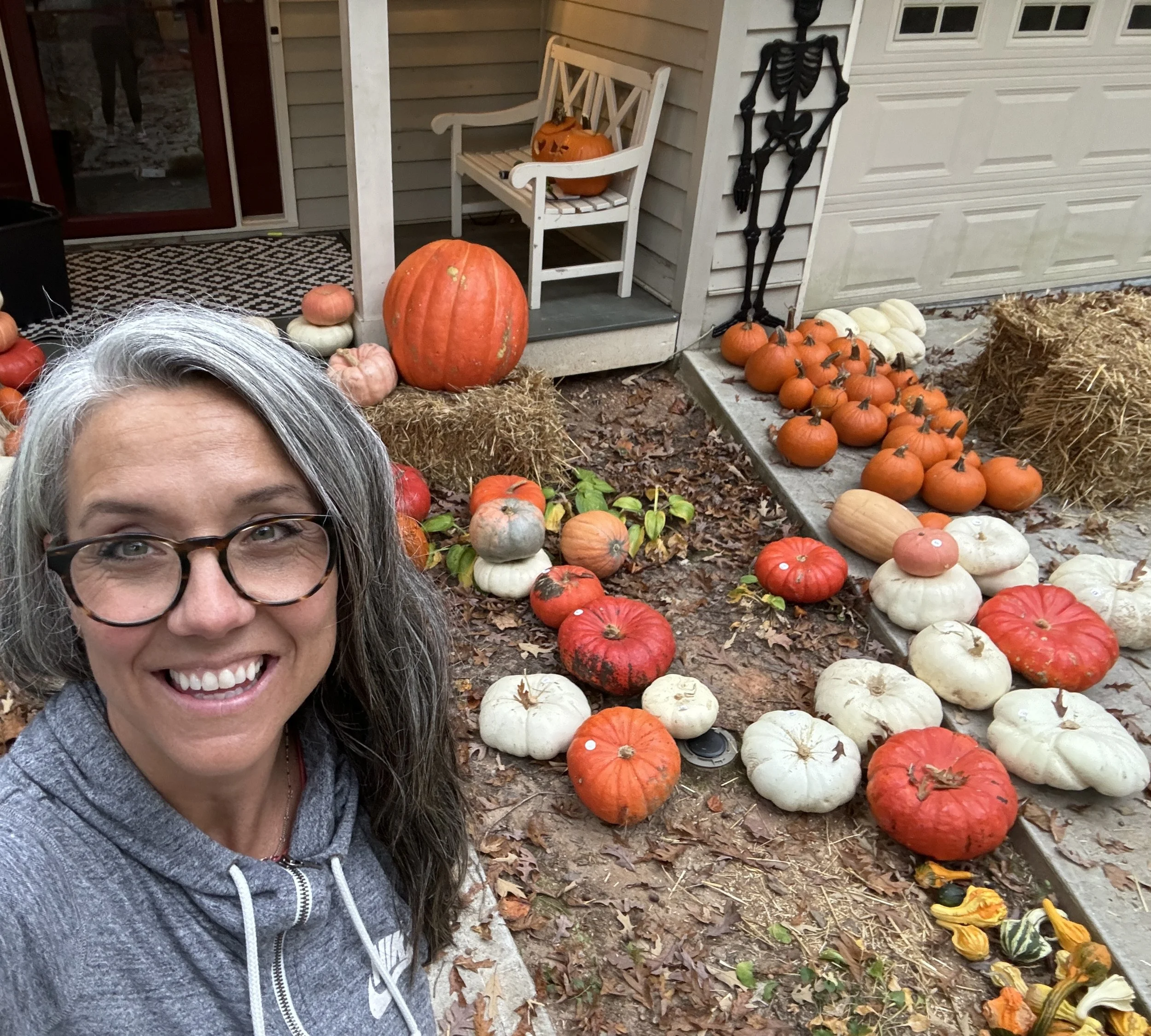 A friendly woman with glasses smiling in a driveway surrounded by autumn pumpkins in various colors and sizes, with Halloween decorations and fall foliage.