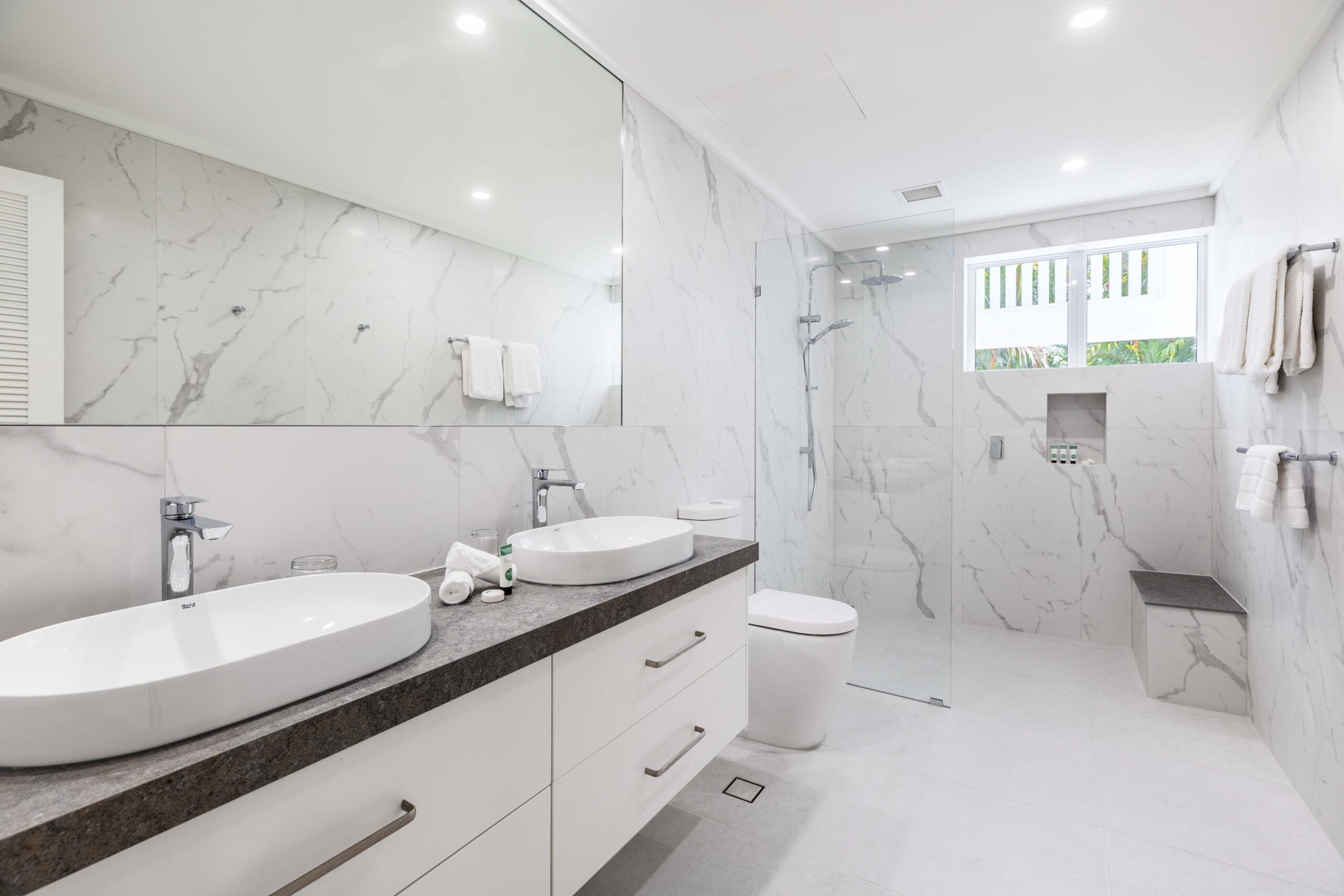 Modern bathroom with white marble walls and floor, double sinks with a dark gray countertop, a large mirror, a window with blinds, towels hanging on racks, a shower area with a glass door, and bathroom toiletries on a recessed shelf.