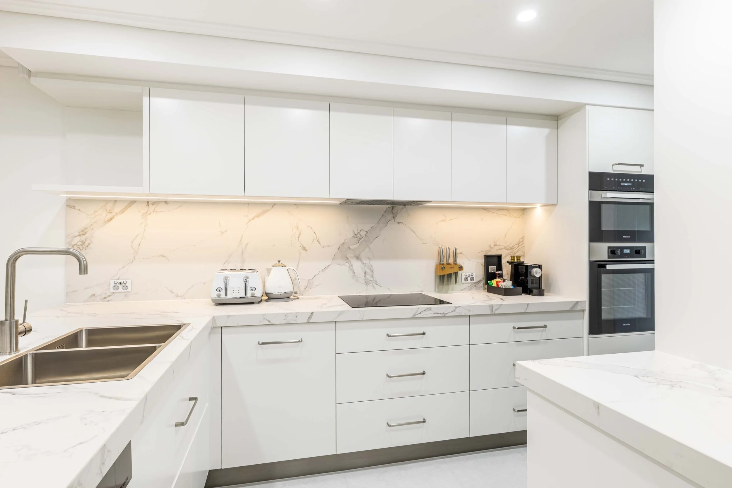Modern kitchen with white cabinets, marble countertops, and a marble backsplash. Includes a double basin sink, toaster, kettle, and built-in oven.