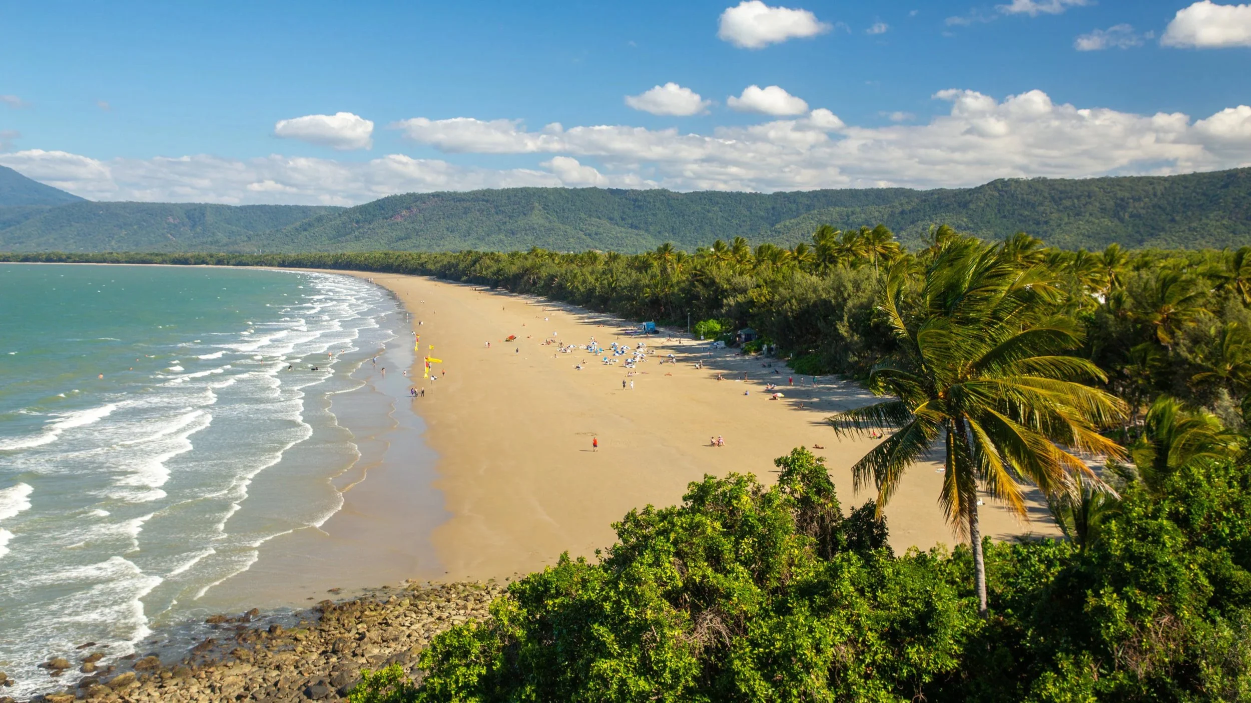 A scenic beach with golden sand, lined with lush green palm trees and surrounded by distant mountains under a partly cloudy sky.