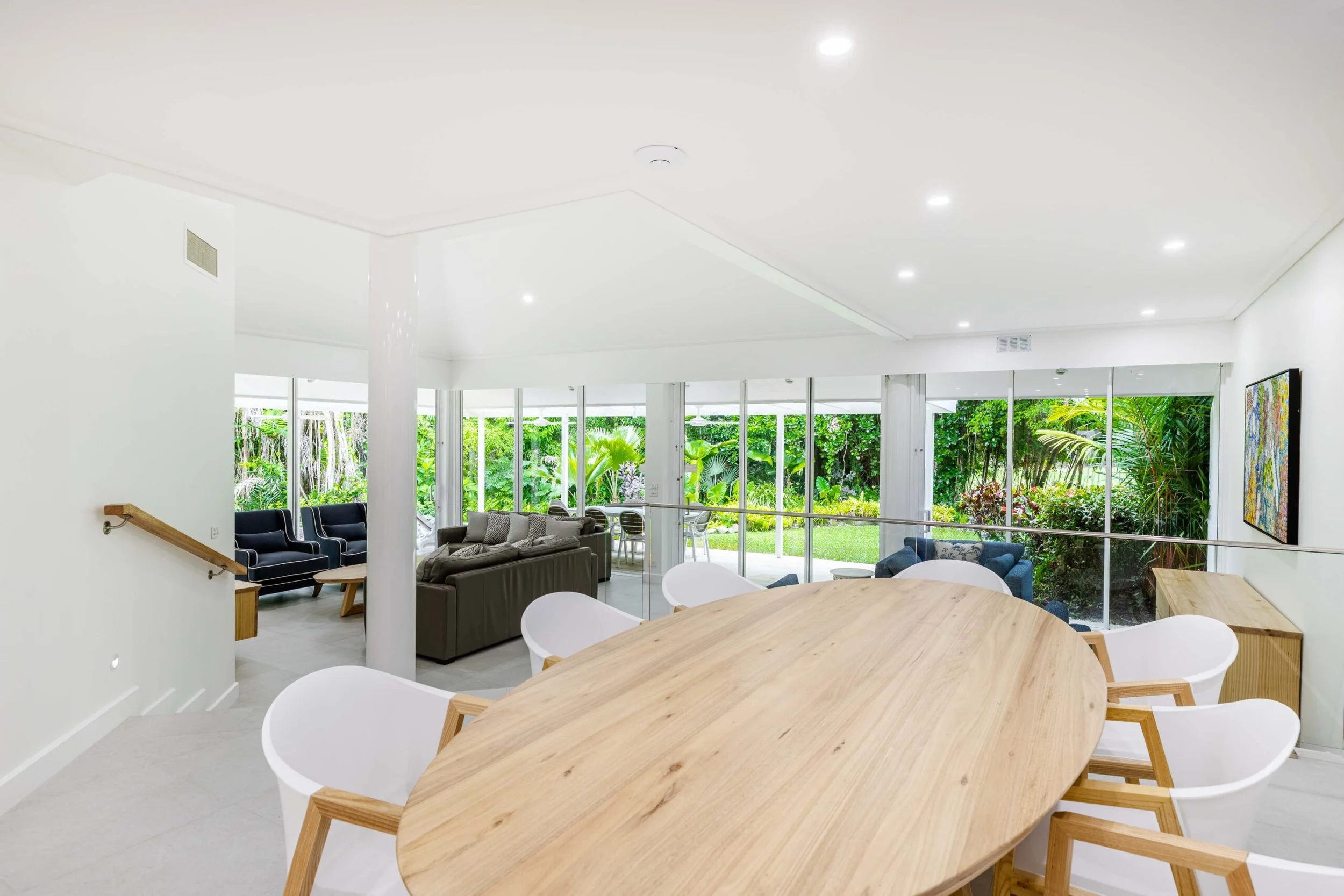 A bright, modern open-plan living and dining area with white walls, large windows overlooking a garden, and a light wood dining table surrounded by white chairs.