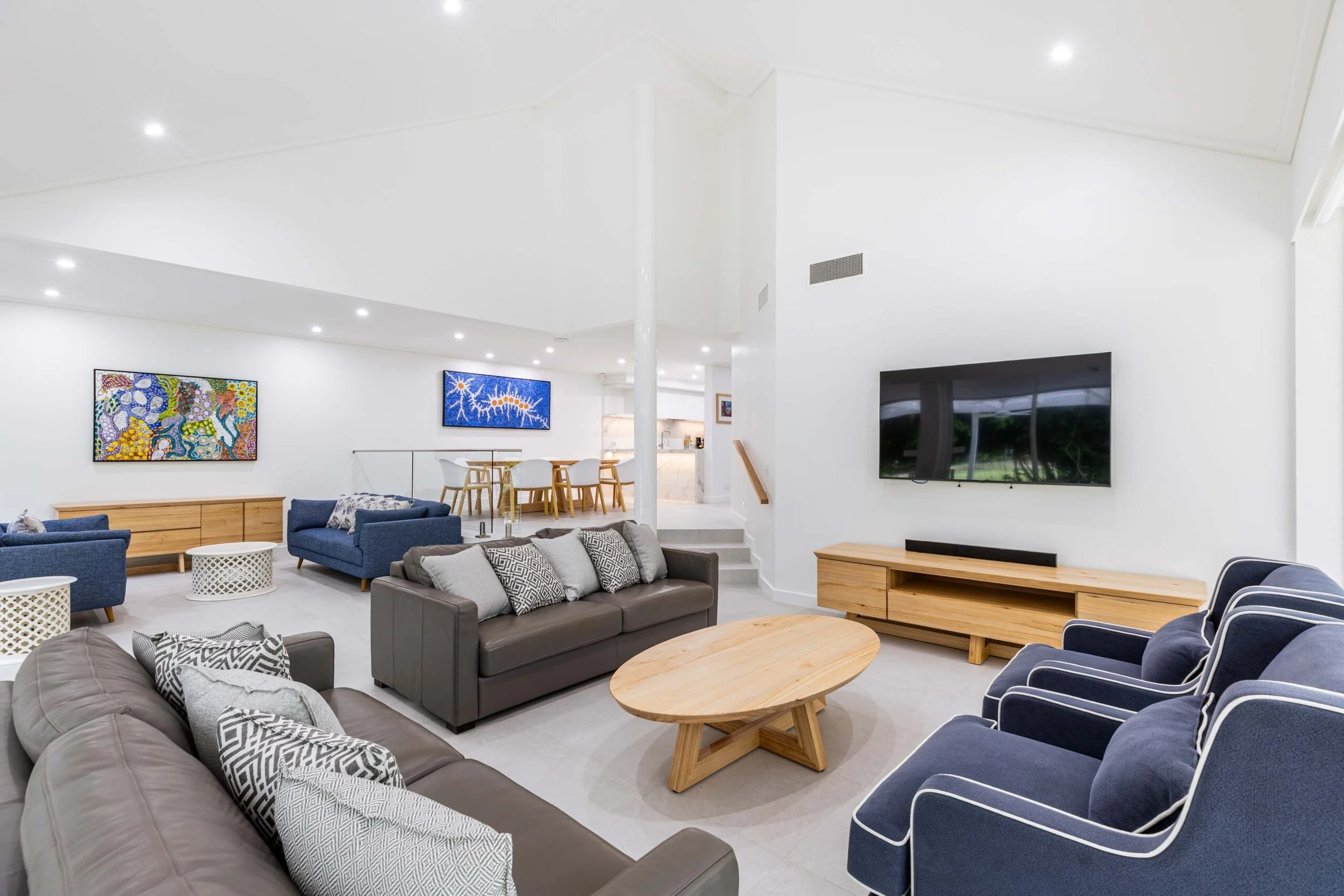 Modern living room with gray, blue, and patterned sofas arranged around wooden coffee tables, colorful artwork on the white walls, a TV mounted on the wall, and an open dining area with chairs in the background.
