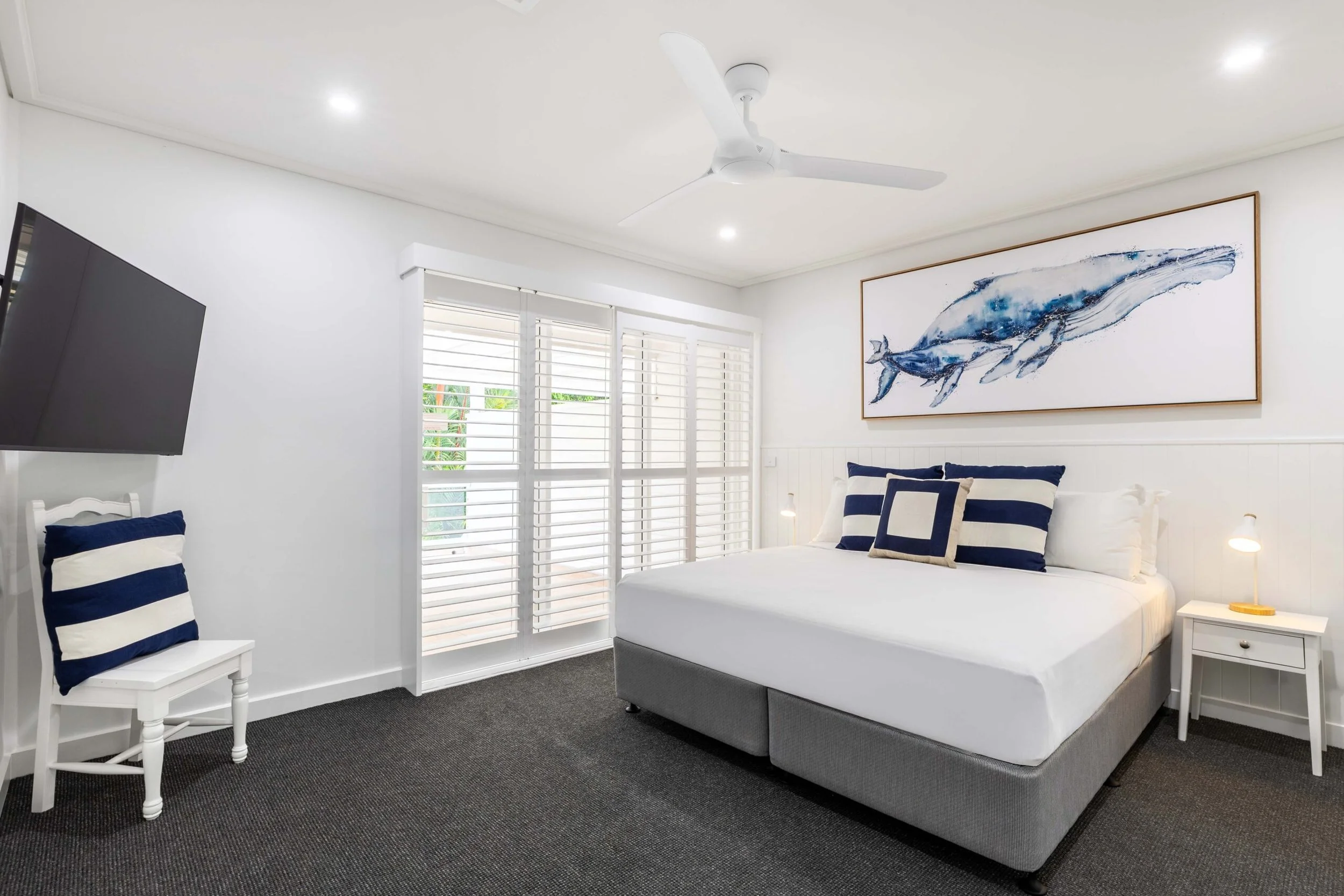 A modern bedroom with white walls, a large framed whale artwork, a white bed with navy striped pillows, white side tables, a flat screen TV, and plantation shutters on the windows.
