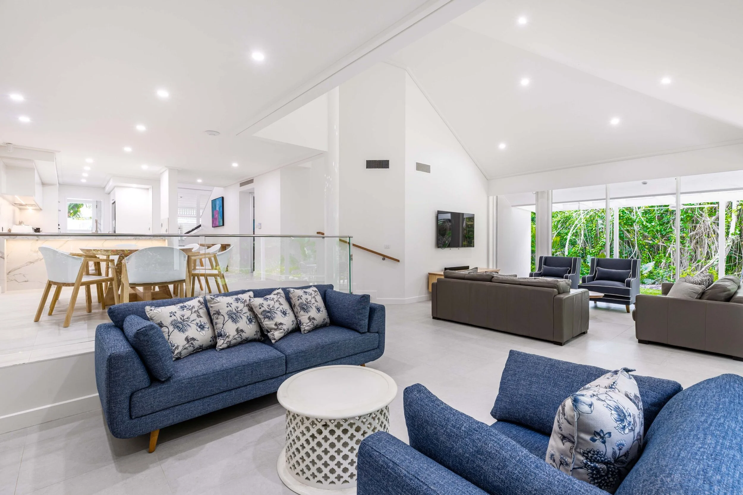 Spacious, modern living room with white walls, large windows showing greenery outside, navy and grey sofas with floral cushions, a white round side table, and a dining area with wooden chairs and a marble dining table in the background.