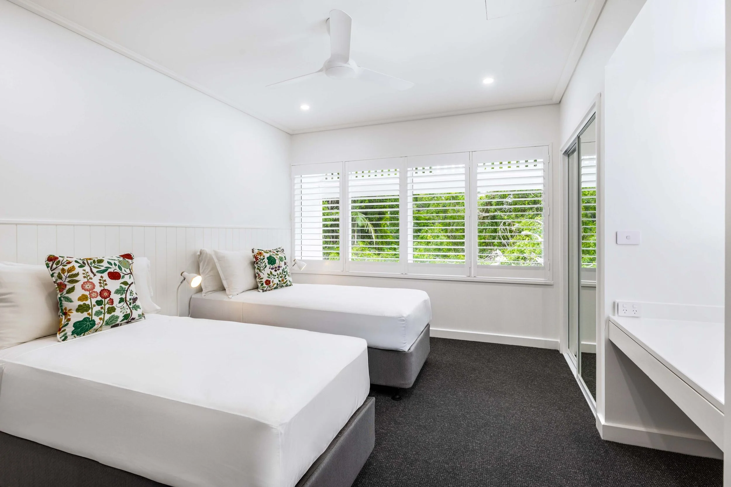 A bright bedroom with white walls, ceiling, and bedding, featuring pillows with colorful floral patterns, a large window with white blinds overlooking green foliage, and a white ceiling fan.