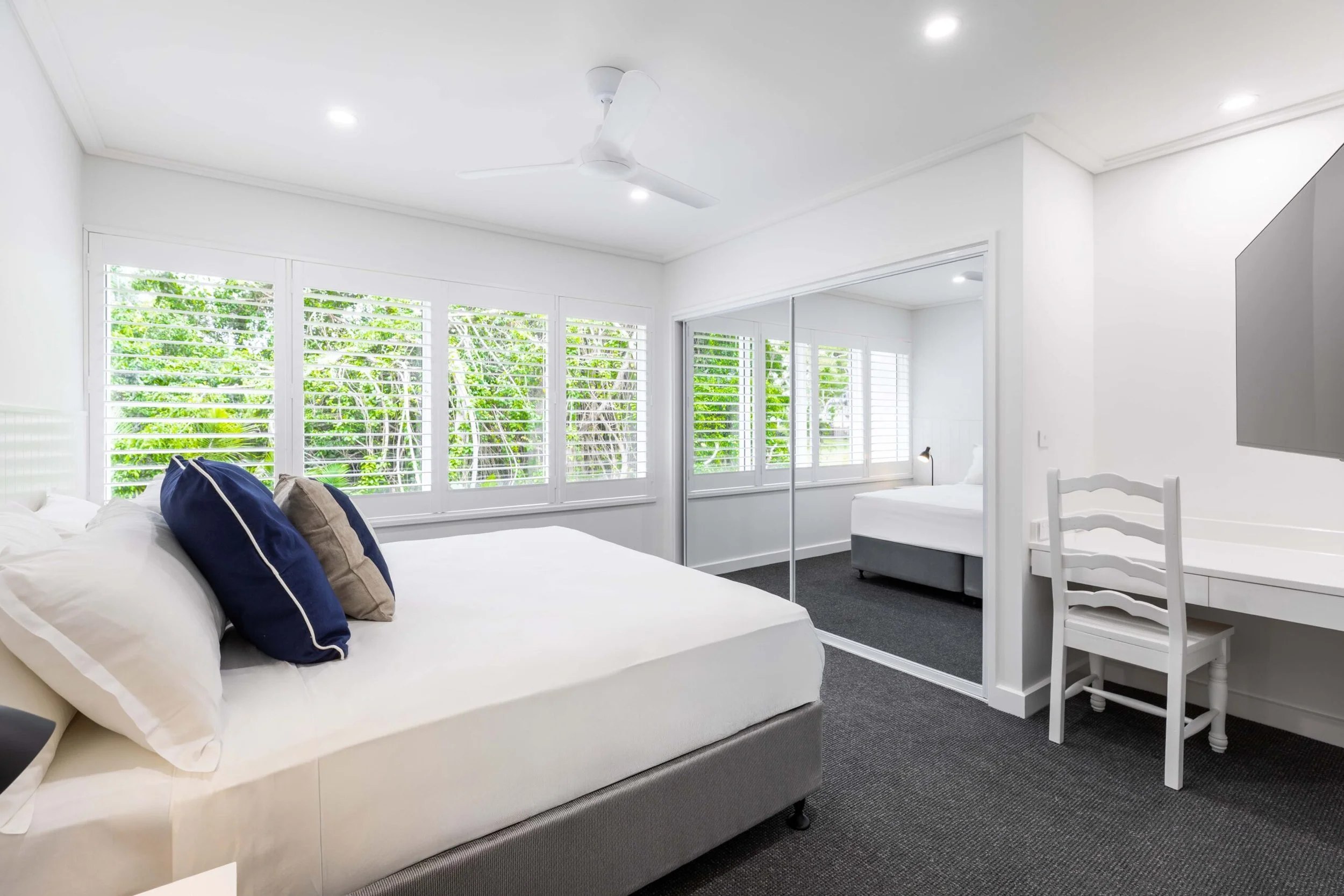 Bright, white-bedroom with large windows showing green trees outside, white bed with navy and beige pillows, white ceiling fan, mirror closet, and small desk with chair.