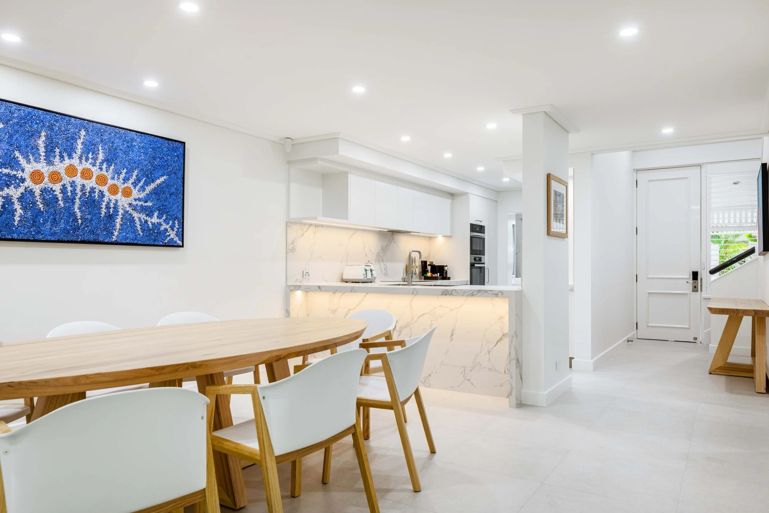 A modern, minimalist kitchen and dining area with white walls and ceiling, a wooden table with white chairs, a marble kitchen island with a sink, built-in oven and cabinets, colorful abstract artwork on the wall, and a window with blinds.