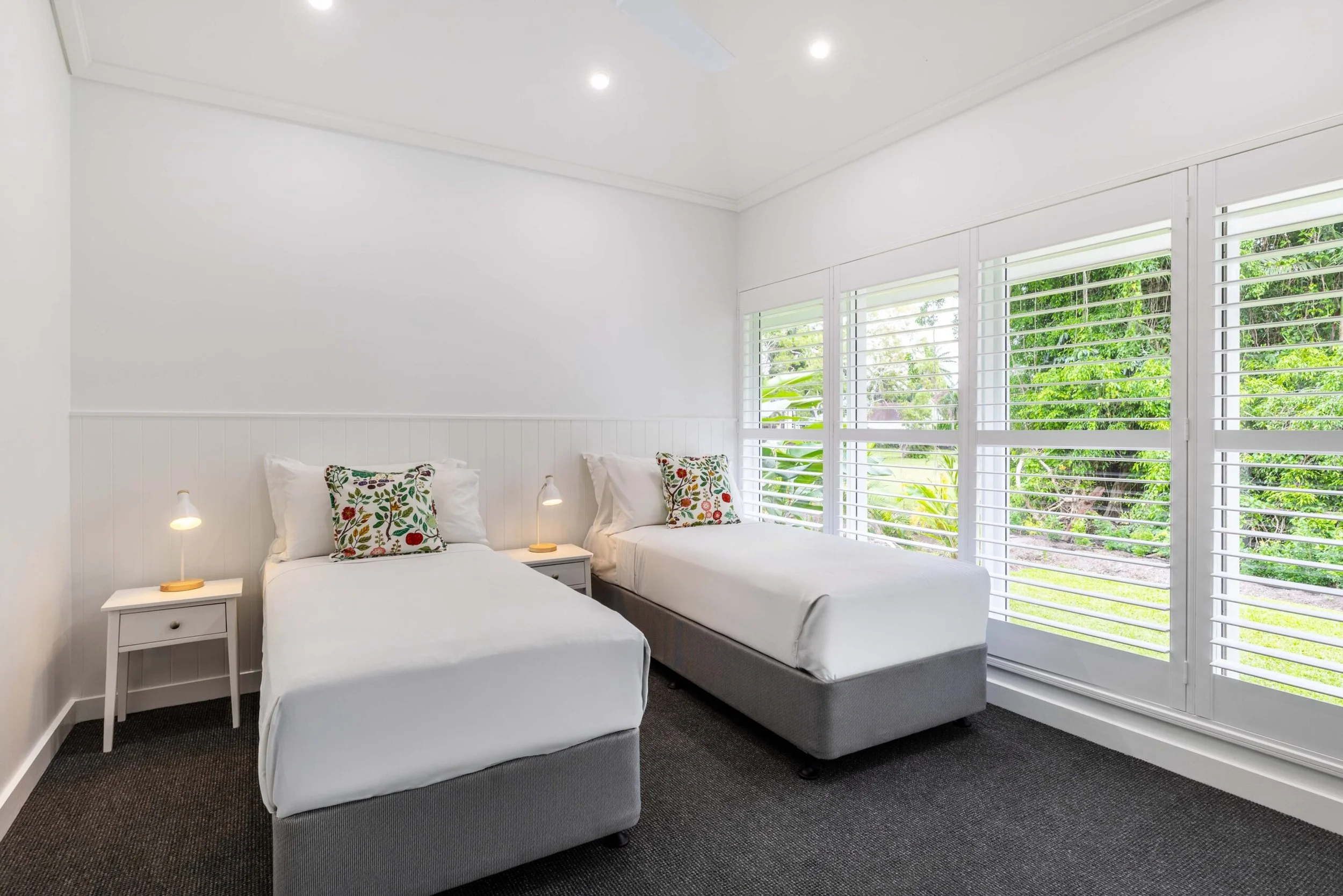 A bedroom with two single beds, white bedding, floral pillows, white nightstands with lamps, large windows with white shutters, green outdoor scenery, and a white ceiling fan.