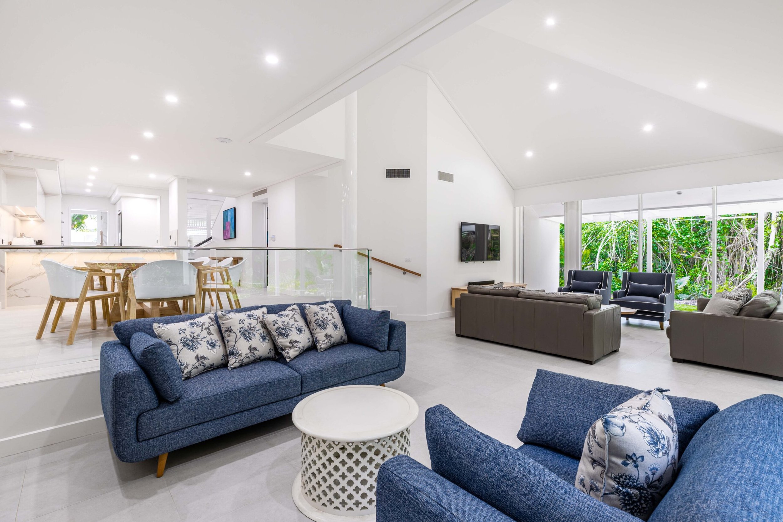 Spacious, modern living room with blue sofas, white round tables, large windows showing green foliage, and a high, white ceiling with recessed lighting.
