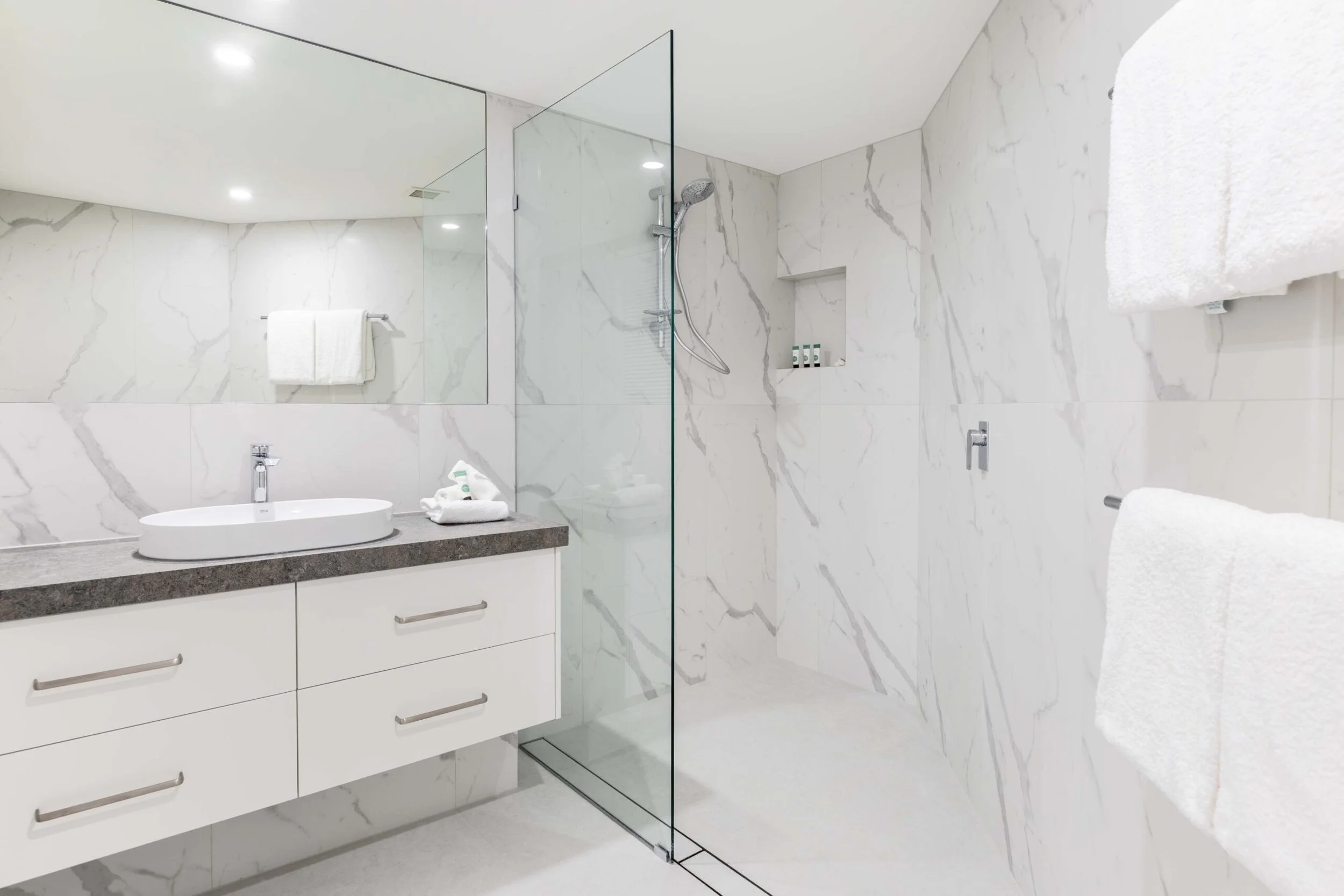 Modern bathroom with white marble walls, a white vanity with a black countertop and an oval sink, and a glass-enclosed shower with a rain showerhead. Towels and toiletries are neatly arranged.