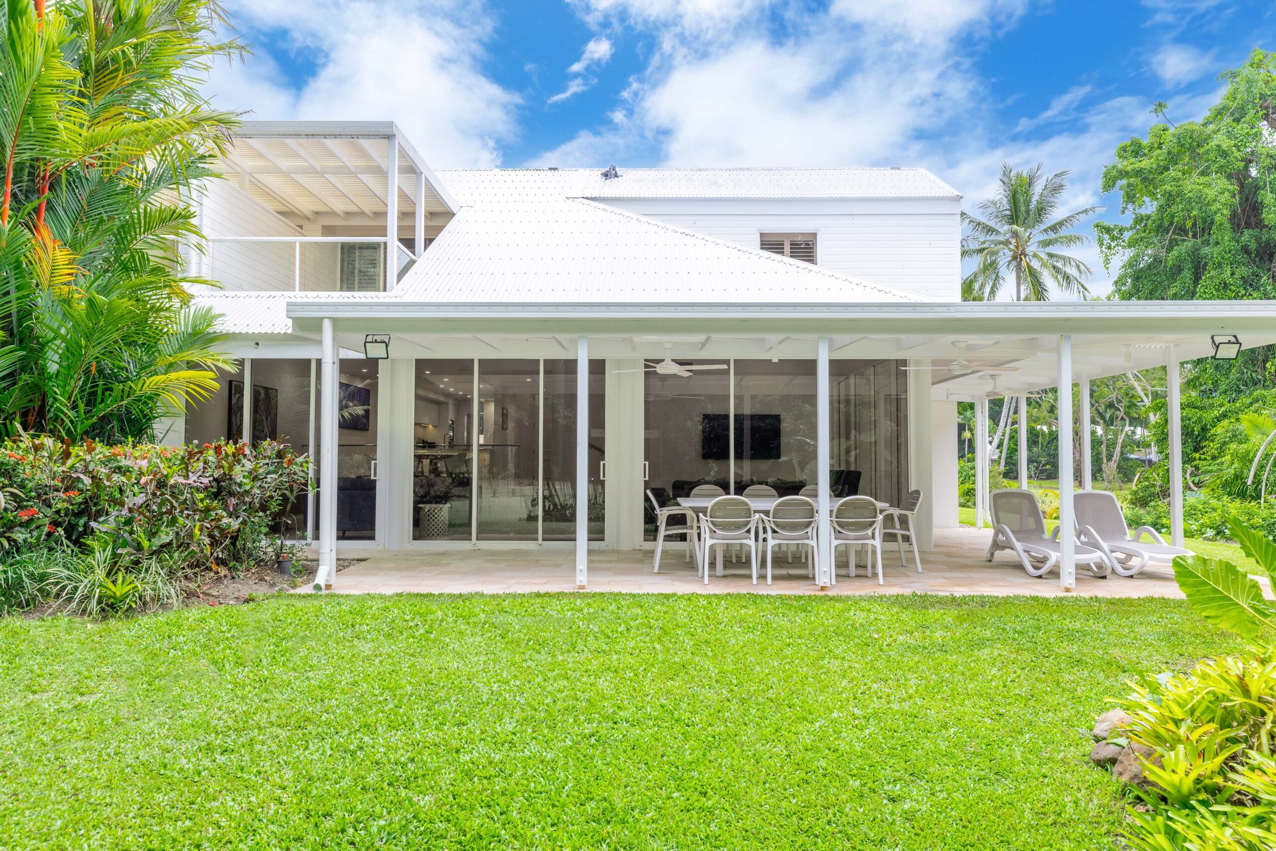 A white modern house with large glass sliding doors, a covered patio with outdoor dining furniture, surrounded by lush green grass and tropical plants, under a blue sky with some clouds.